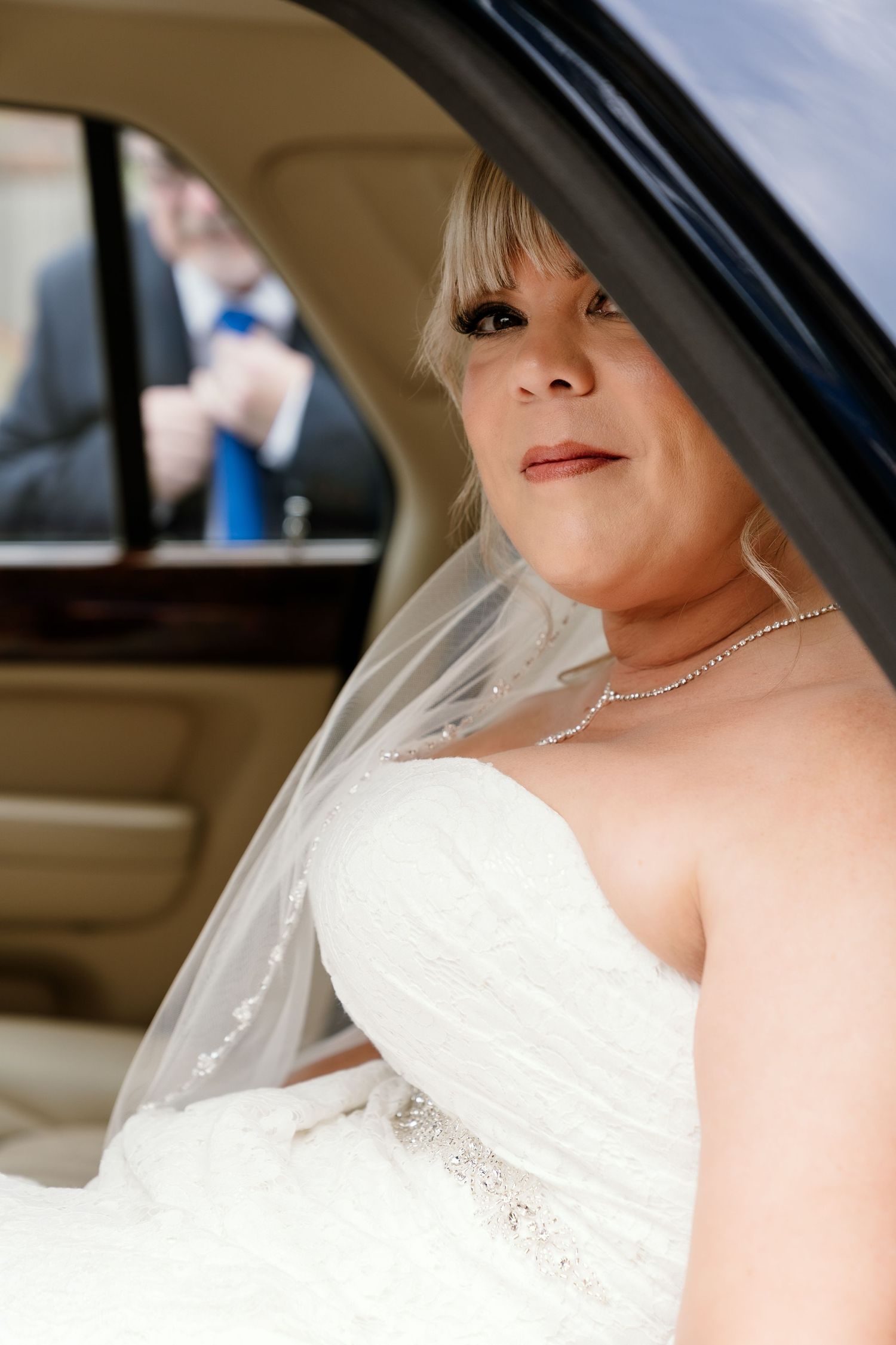 A bride wearing a white strapless wedding gown and crystal necklace sits in a car on her wedding day.