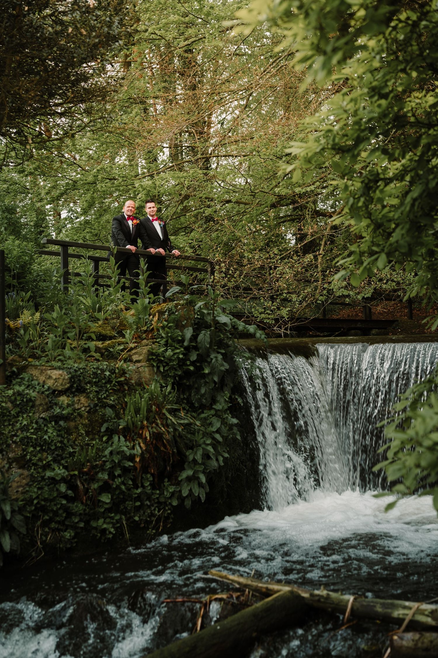Wedding couple stands on wooden bridge overlooking scenic waterfall surrounded by lush greenery.