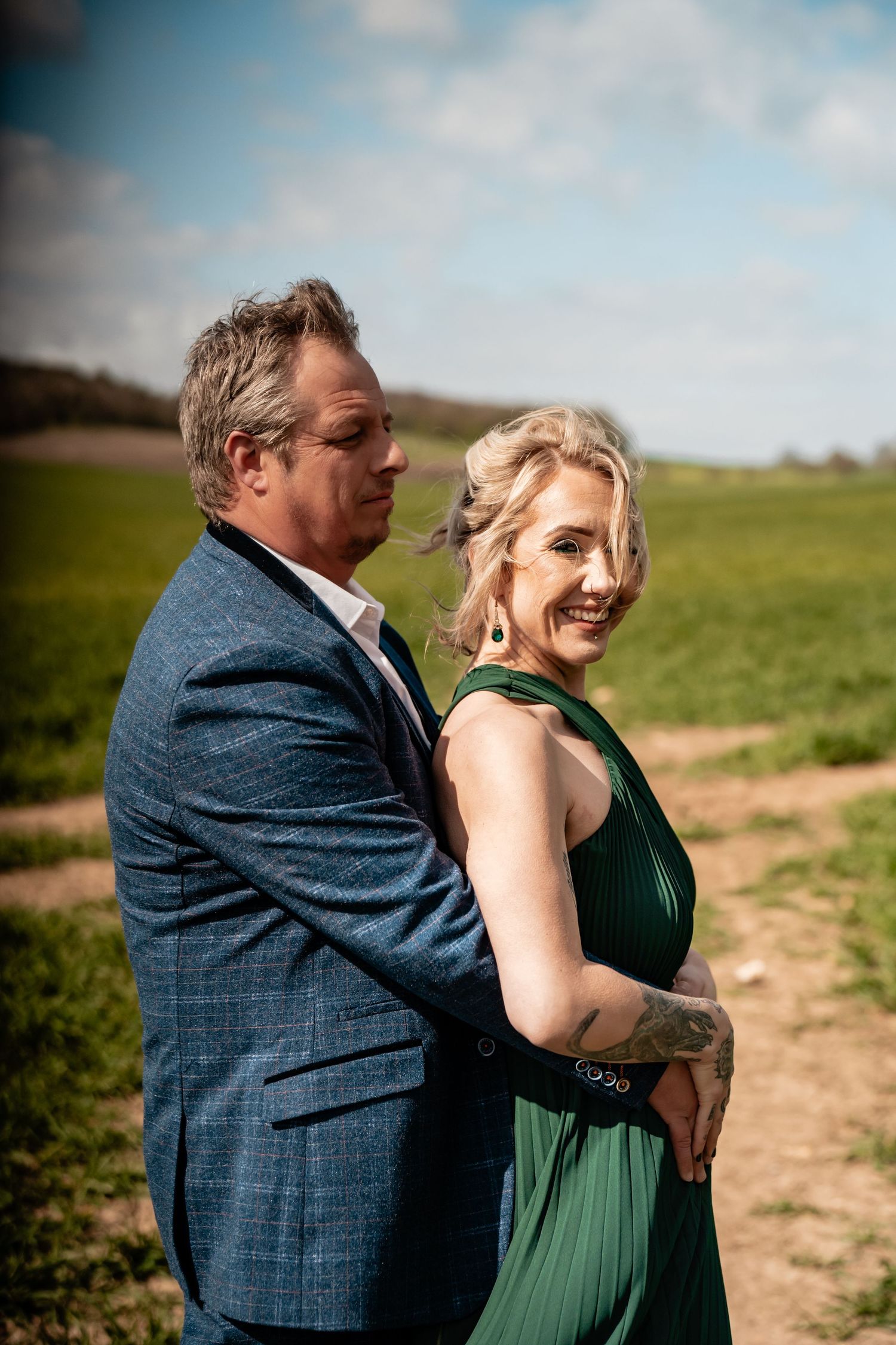 A romantic outdoor portrait of a couple embracing and laughing together in formal attire against a rural backdrop.