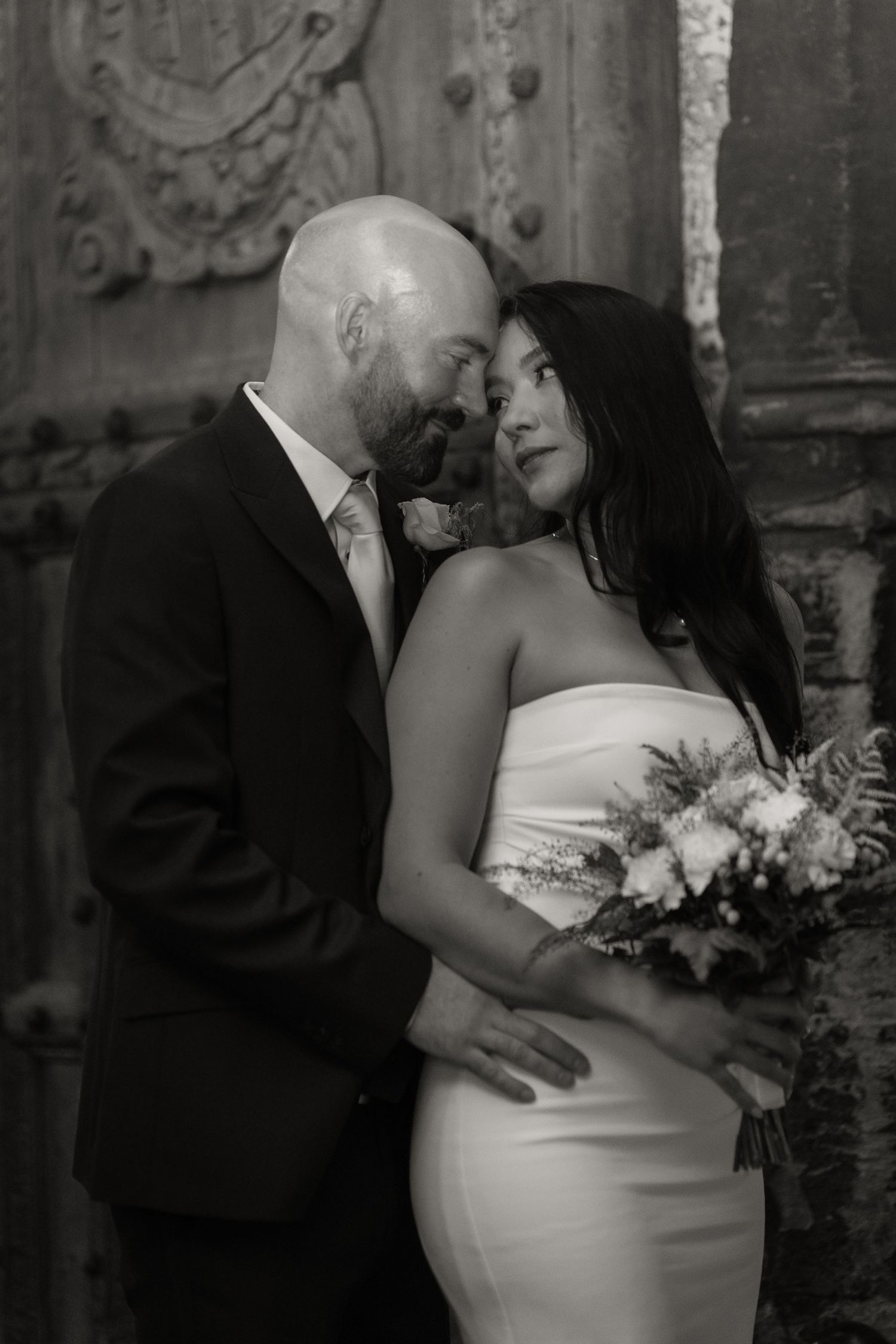 An intimate black and white portrait of newlyweds embracing against an ornate architectural backdrop.