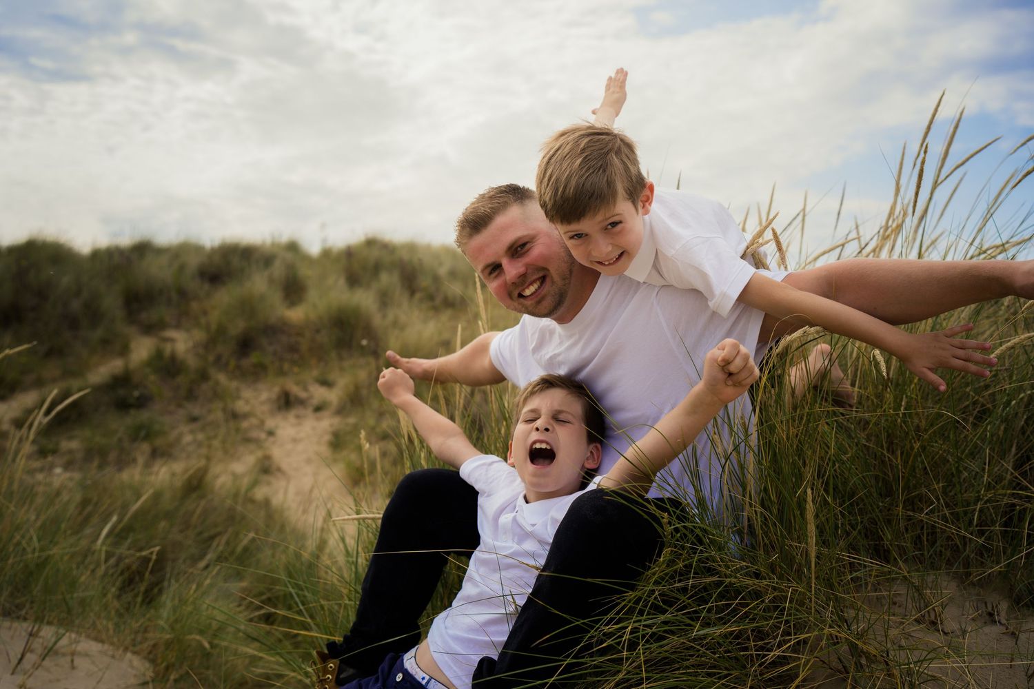 Playful family interaction on grassy coastal dunes during cloudy day.
