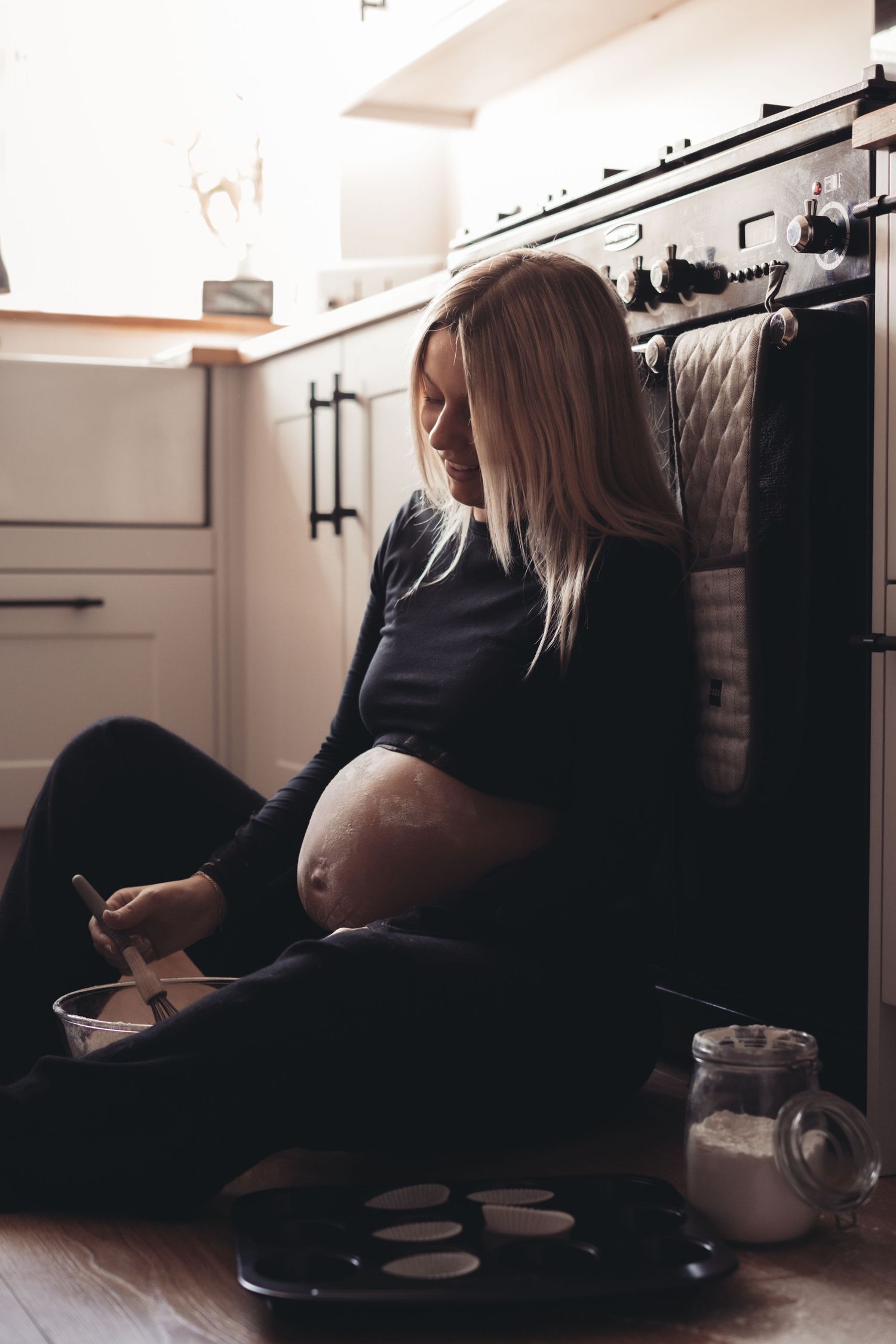 Side view of pregnant woman sitting on kitchen floor with mixing bowls.