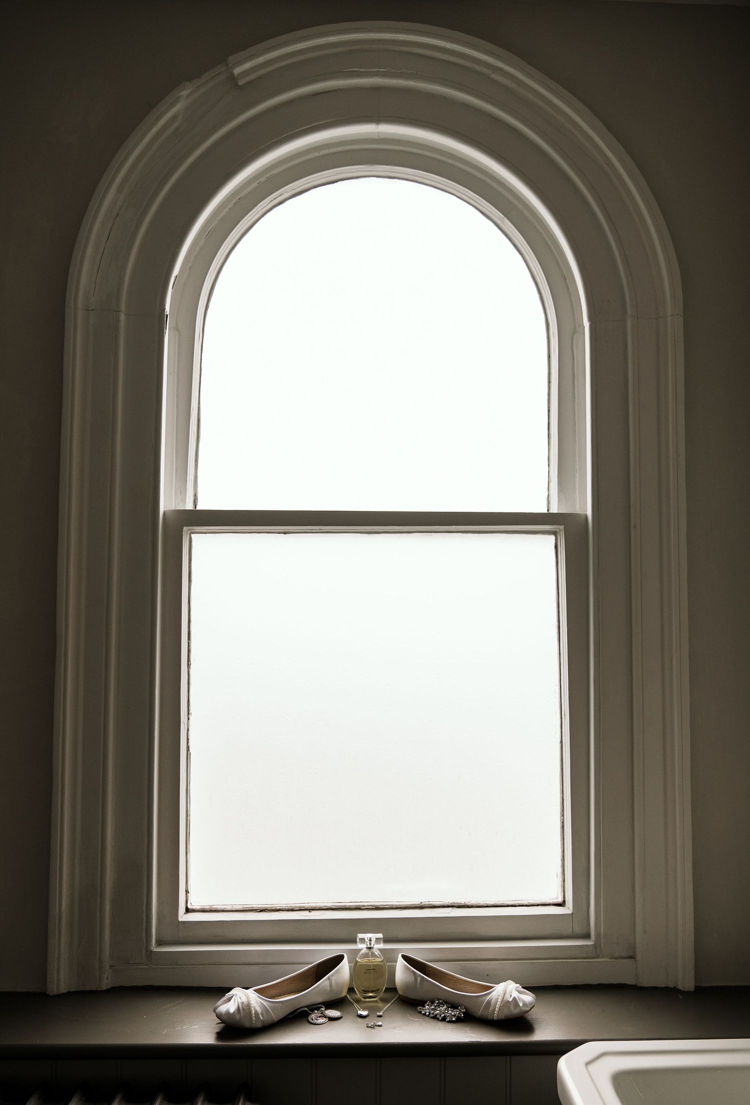 Wedding shoes and perfume bottle displayed in arched window silhouette.