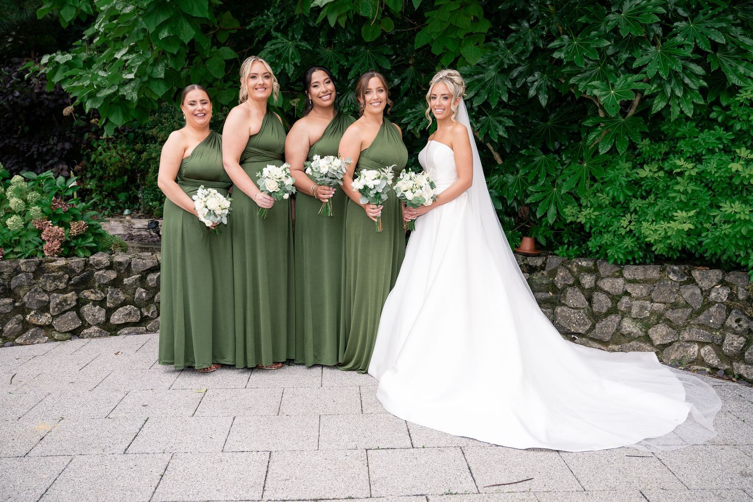 Bridal party in elegant green dresses poses with bride in white gown outdoors.