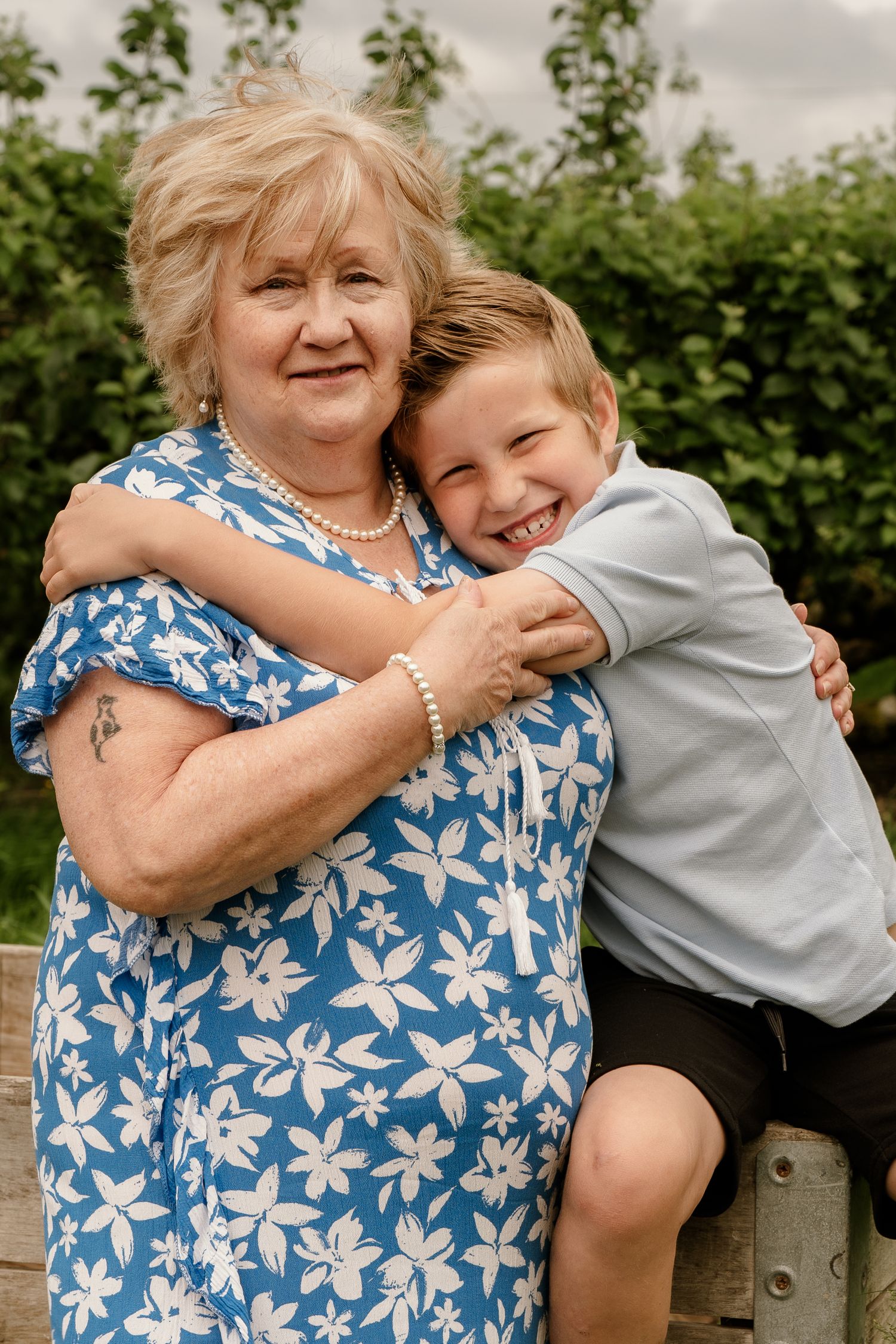 Person in blue floral dress embraces youth in gray shirt in outdoor garden setting.