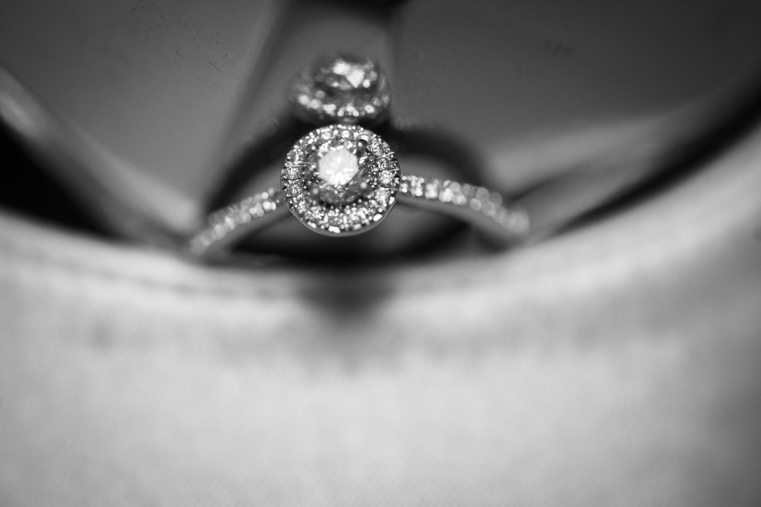 Close-up black and white macro shot of a diamond engagement ring with a halo setting resting on a soft surface.