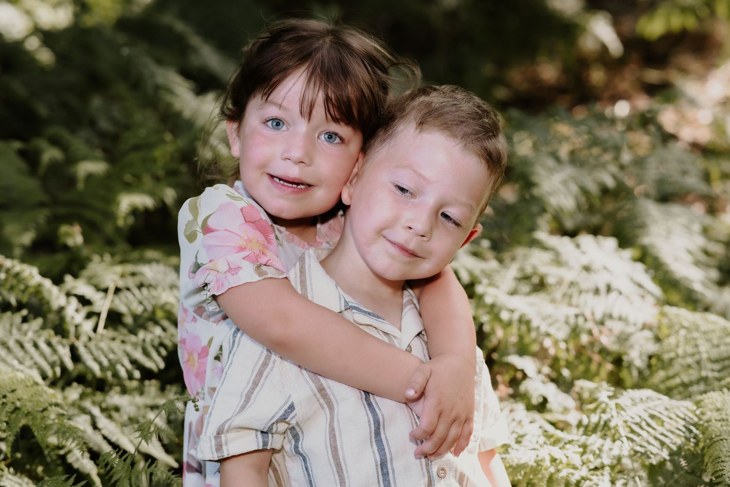 Two children hugging and smiling among green ferns in a woodland setting.
