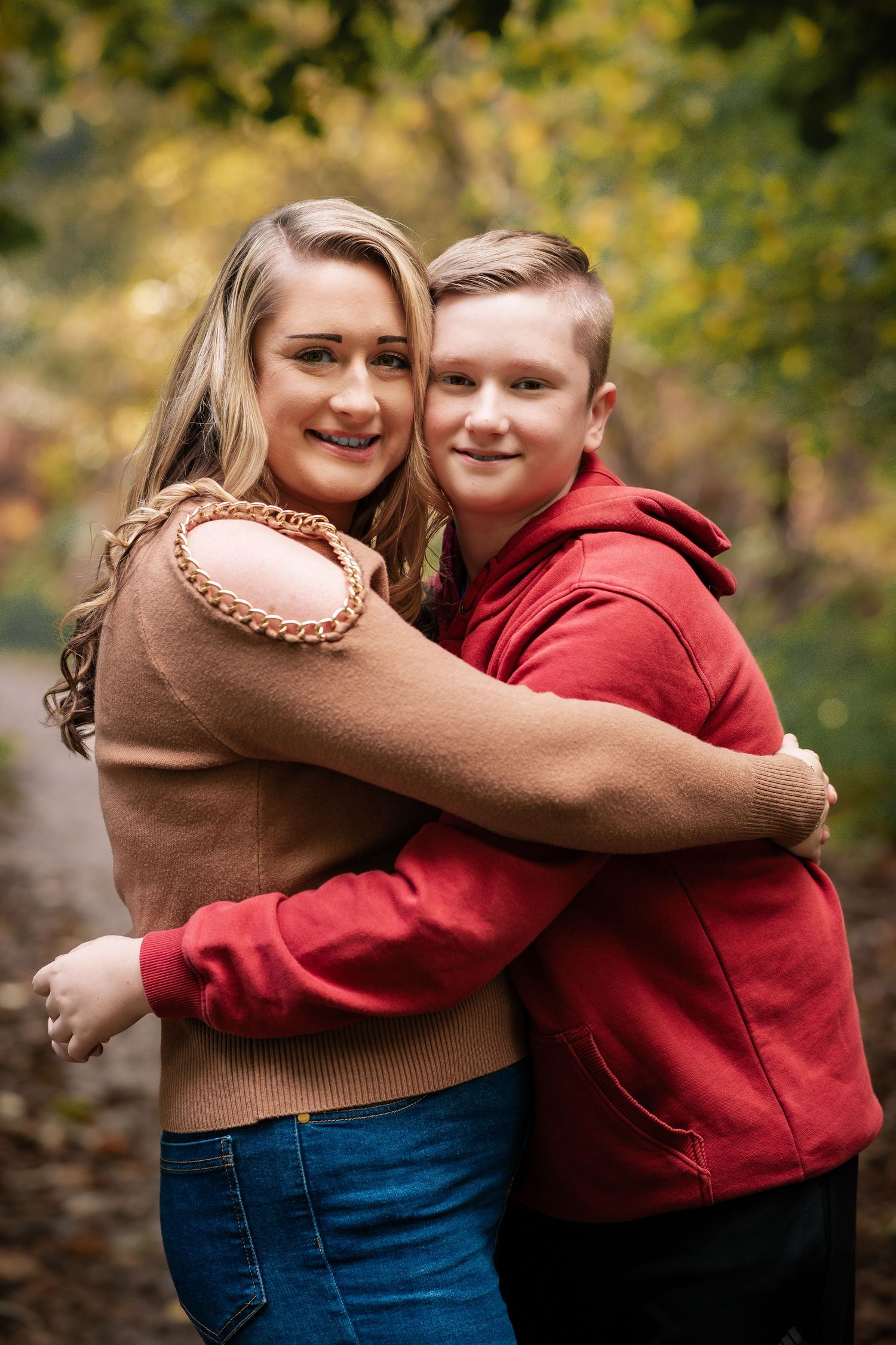 Two people in brown and red sweaters share an embrace on an autumn trail.