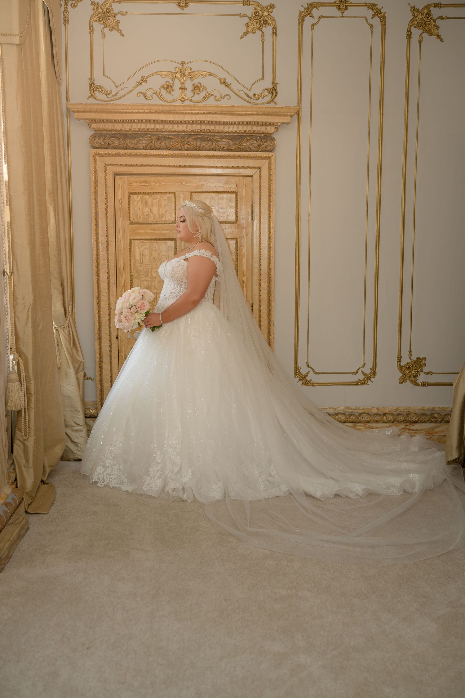 A bride in a flowing ball gown poses in front of an ornate gold-detailed wall.