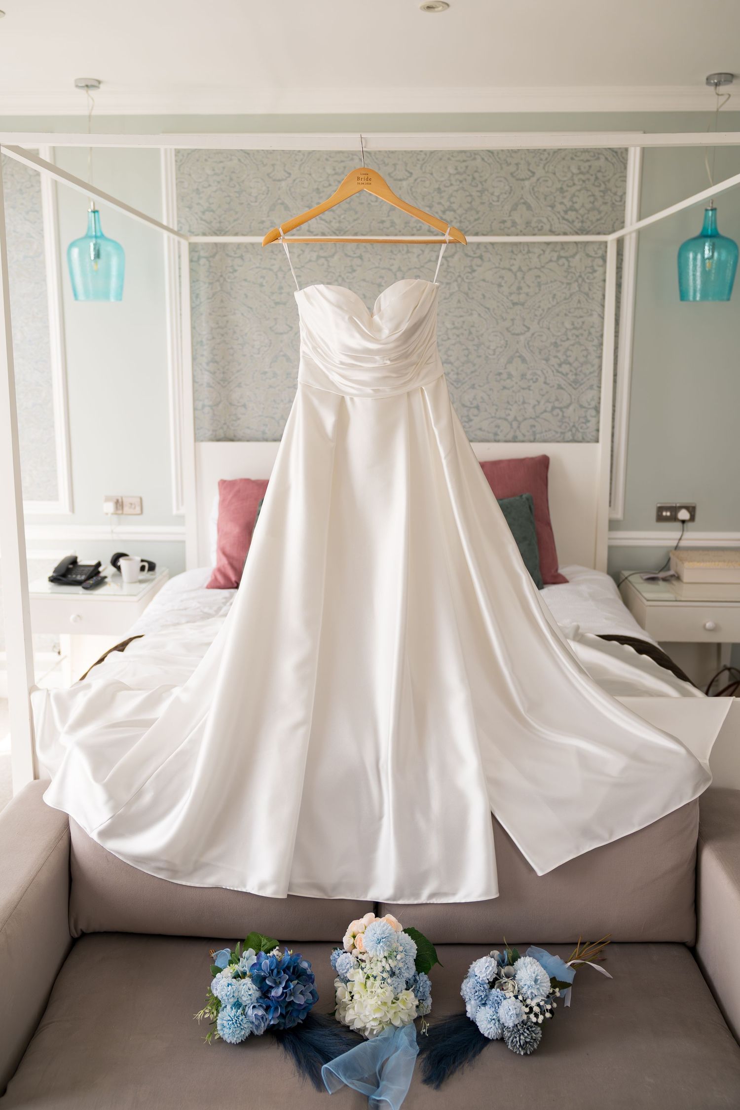Elegant white strapless wedding gown hanging with blue floral bouquets displayed beneath.