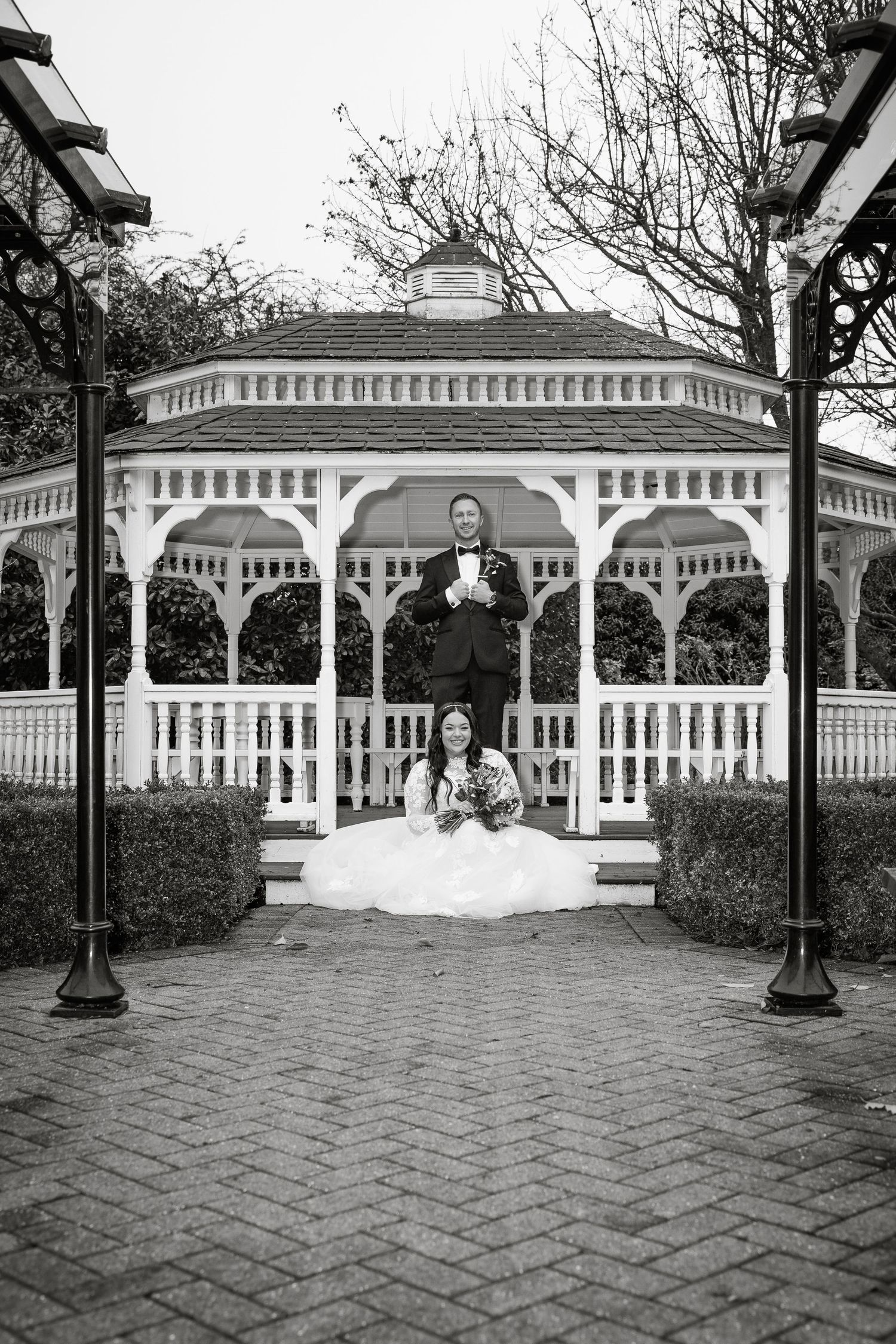 A black and white wedding portrait featuring a classic white gazebo and brick pathway.