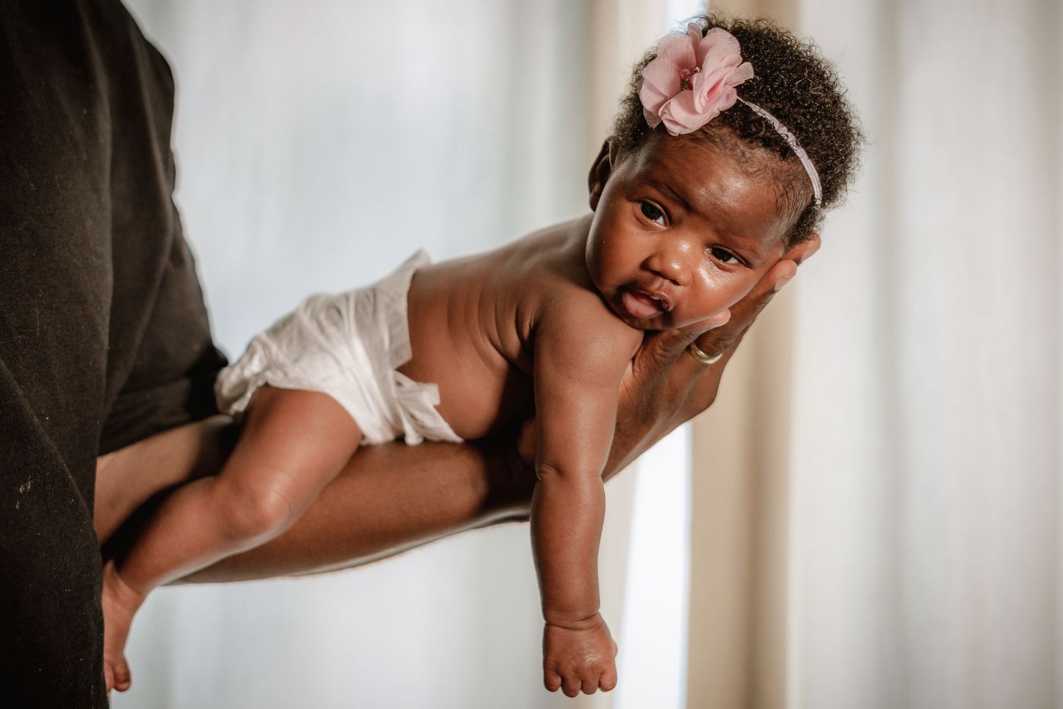 Baby in white outfit with pink bow being held against window light.