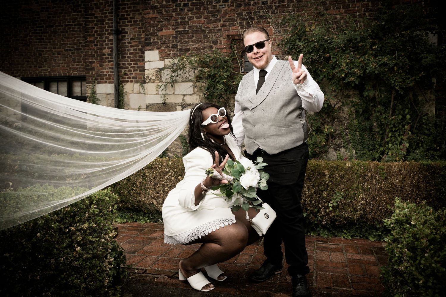 Creative wedding portrait with flowing veil against brick wall background.
