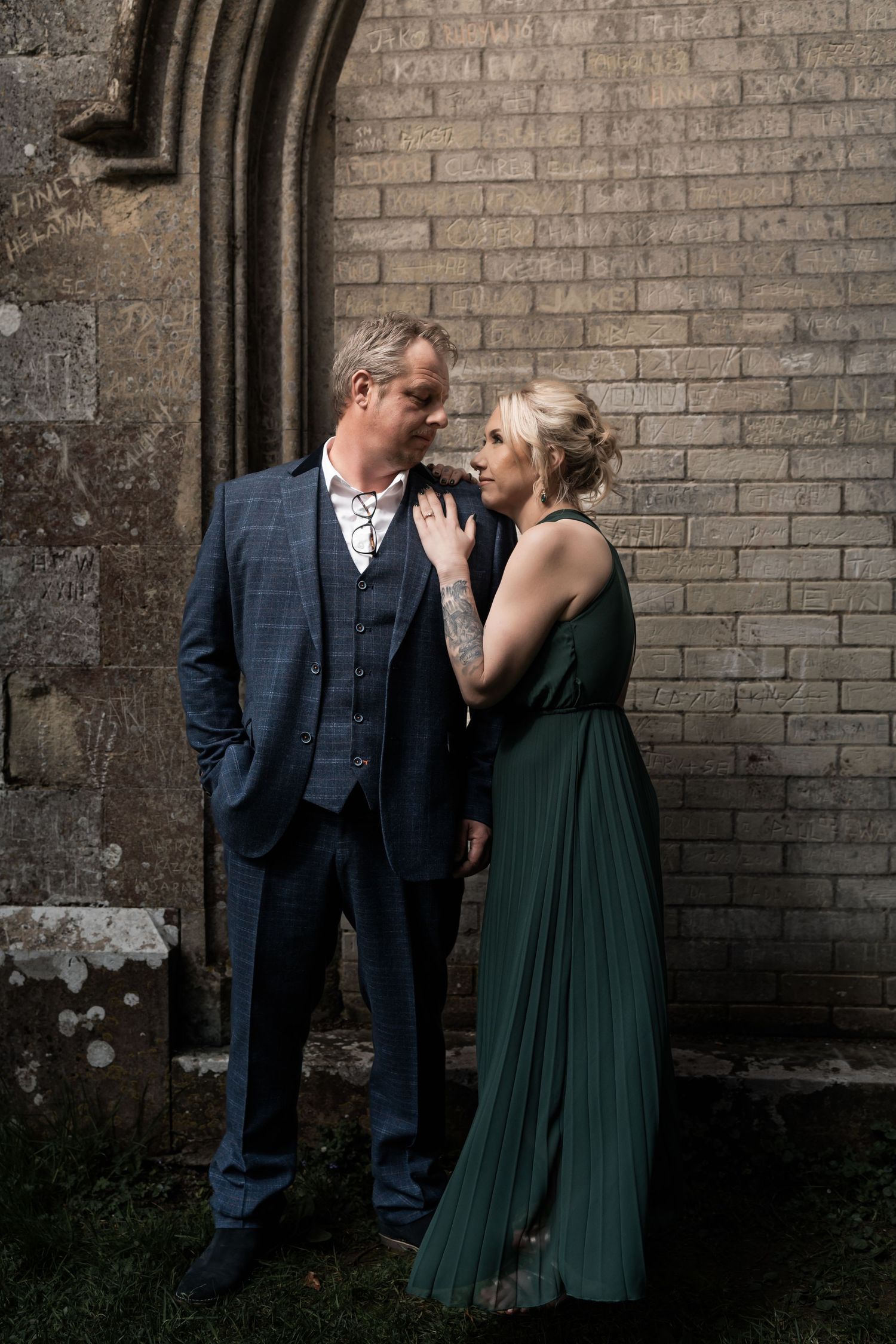 A couple in formal attire share an intimate moment against a brick wall, with her wearing an elegant emerald green gown.