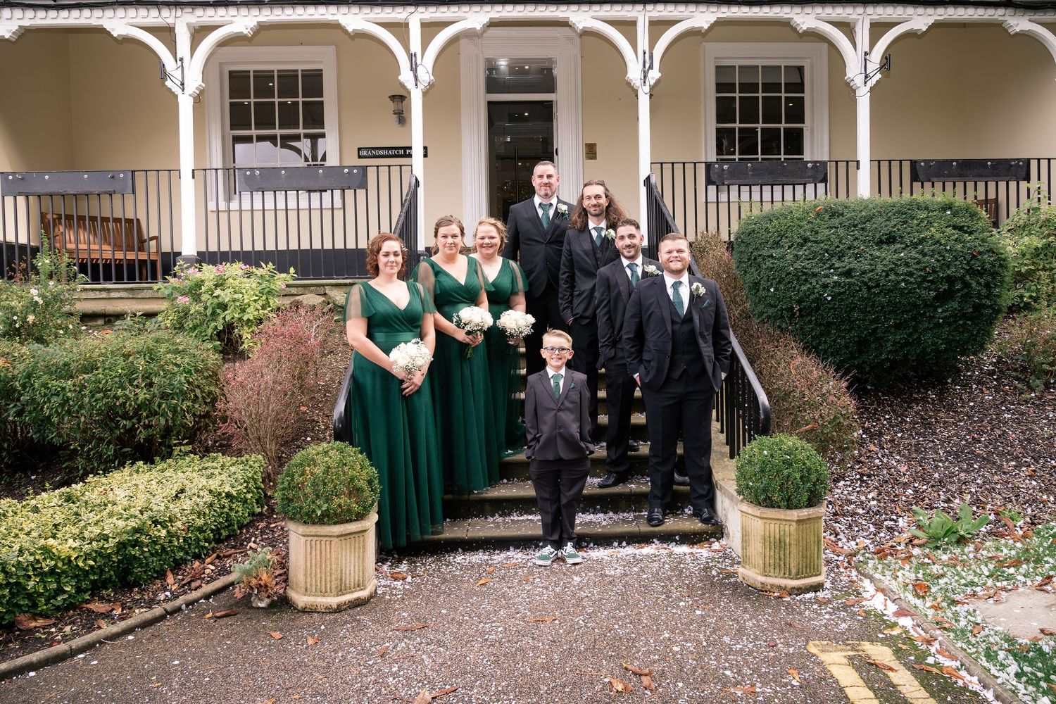 Wedding party in dark green dresses and suits pose on brick building steps.