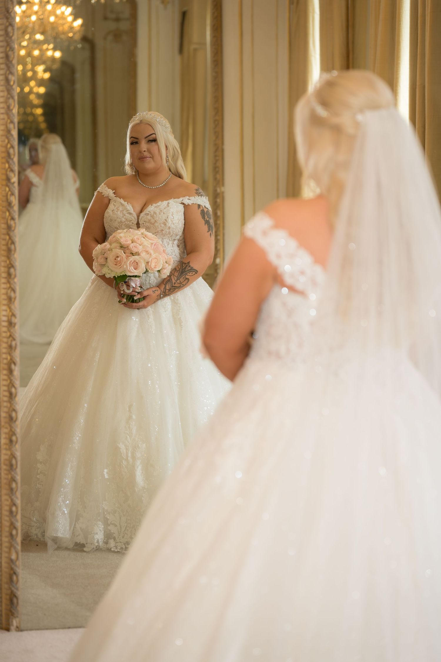 A bride in a beautiful ball gown looks at her reflection in an ornate mirror.