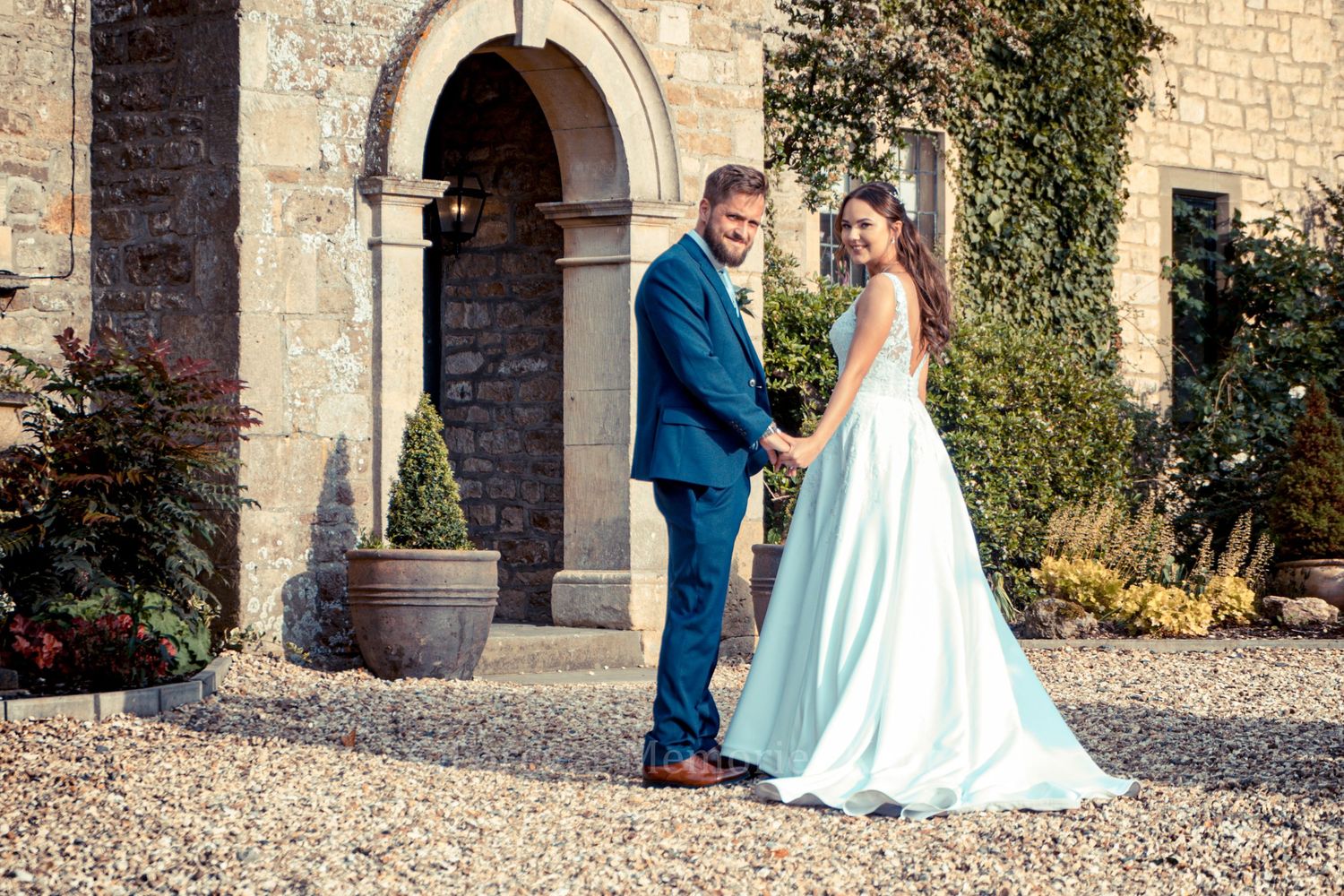 A couple stands in front of a stone archway entrance wearing wedding attire on their special day.