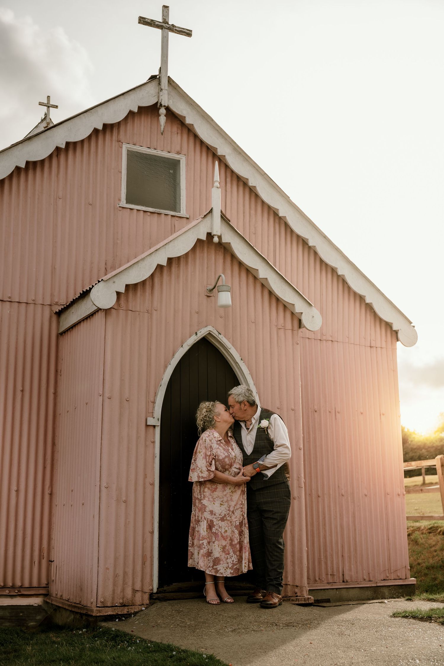 A pink wooden church with crosses stands in warm sunset light with two people embracing in the arched doorway.