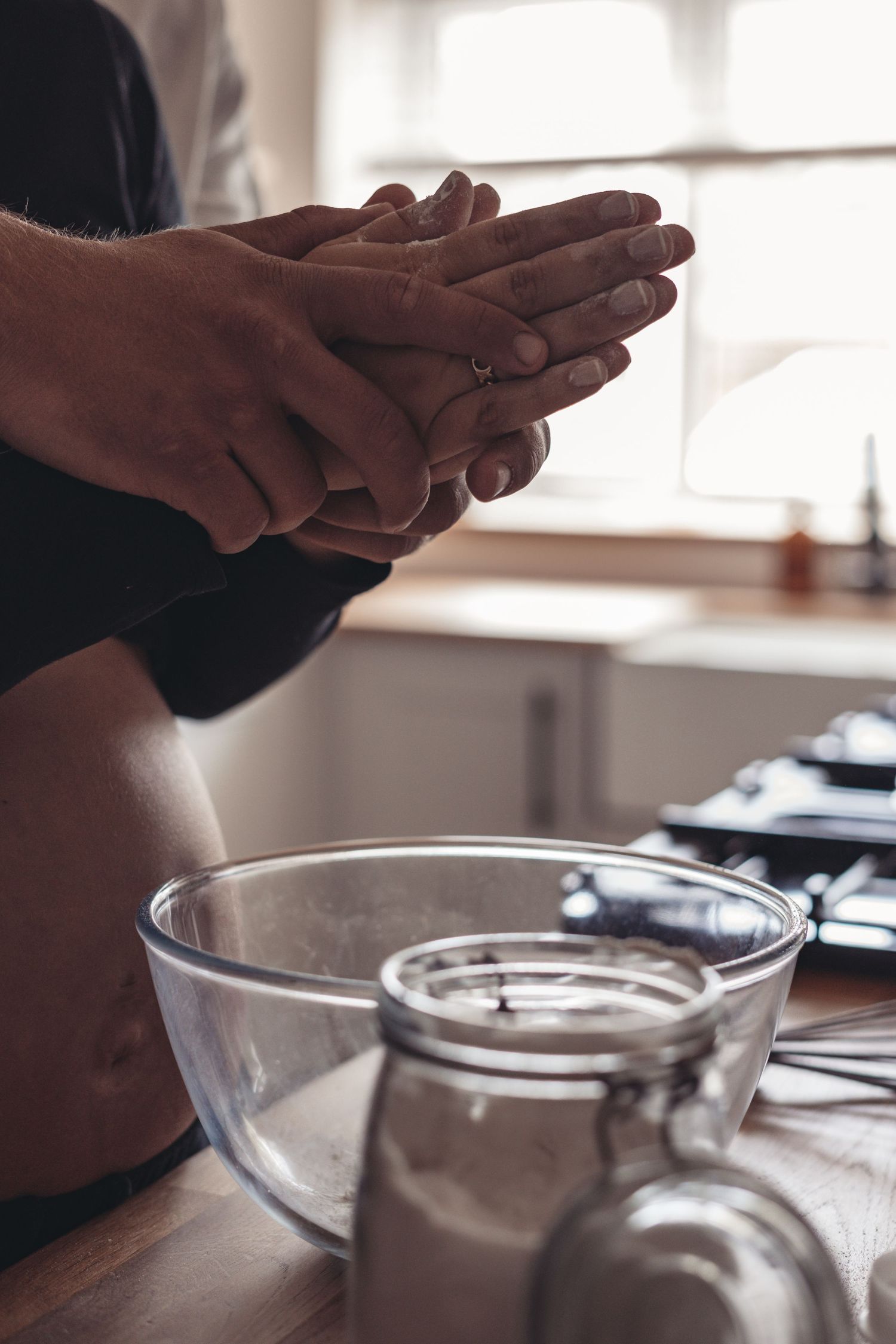 Hands intertwined over glass mixing bowl in sunlit kitchen.