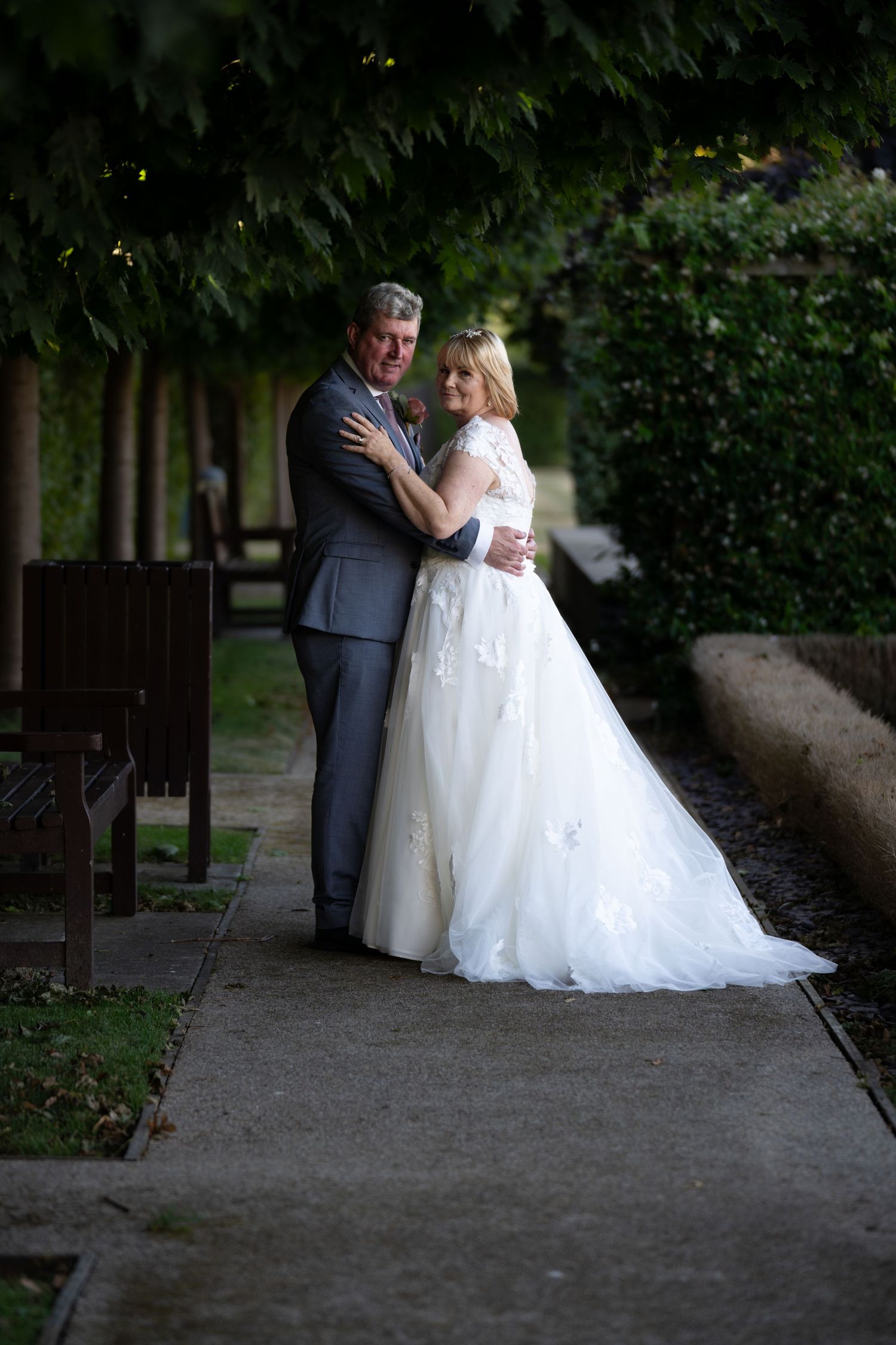 Wedding couple shares intimate moment along tree-lined garden path.