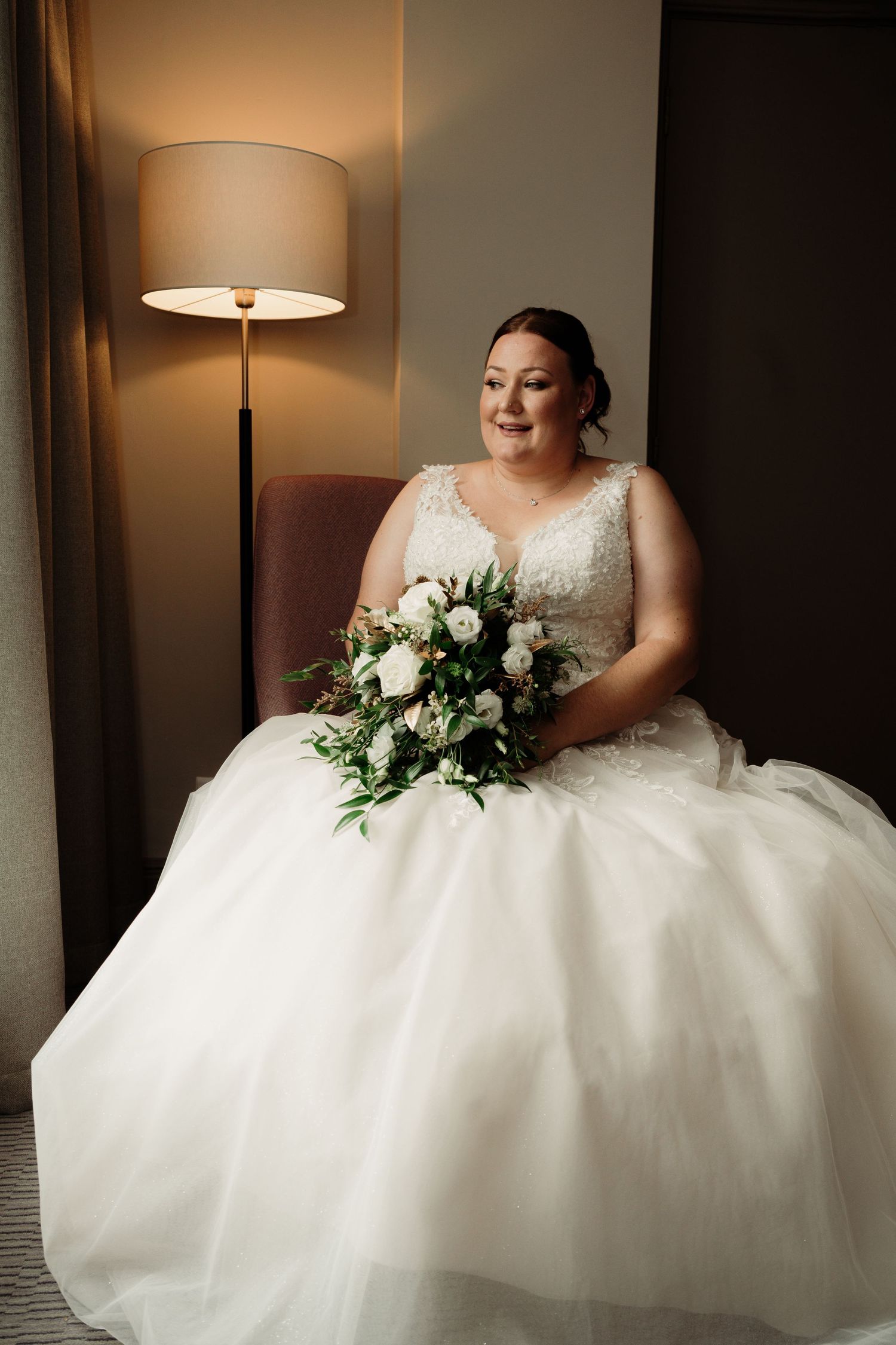 Elegant bride in white lace gown sits with bouquet by warm-lit lamp.