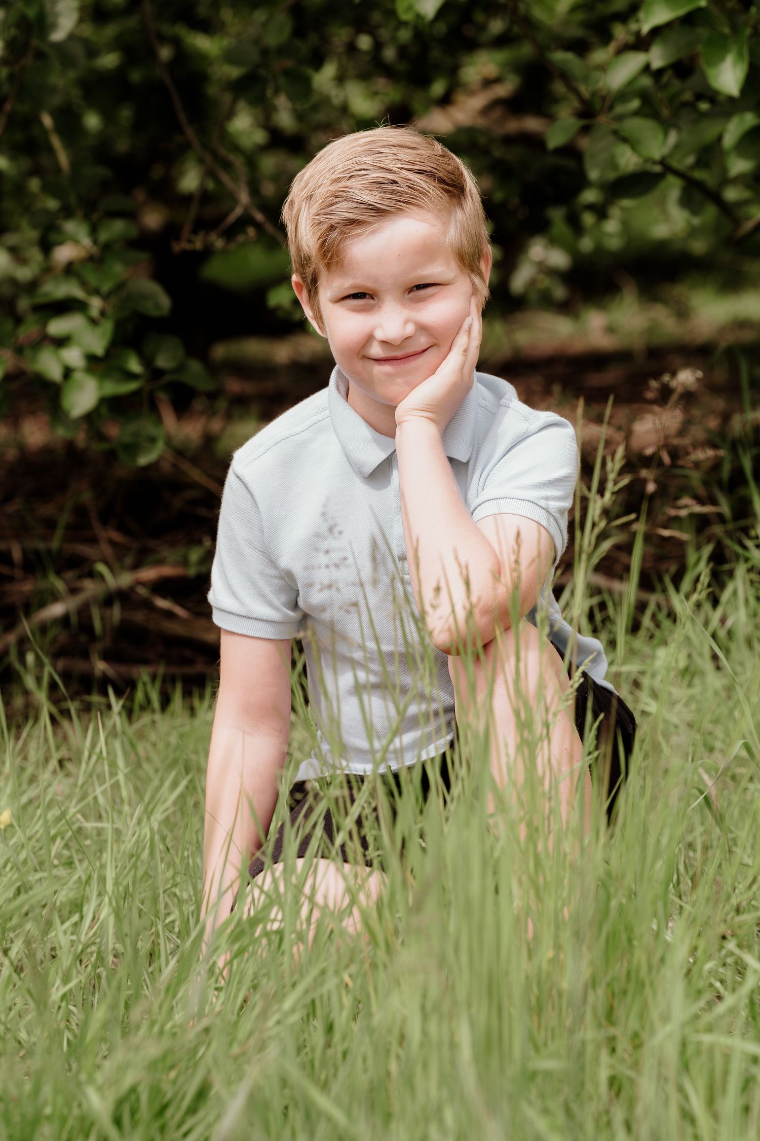 Person in white polo shirt poses in tall grass with hand resting on chin.