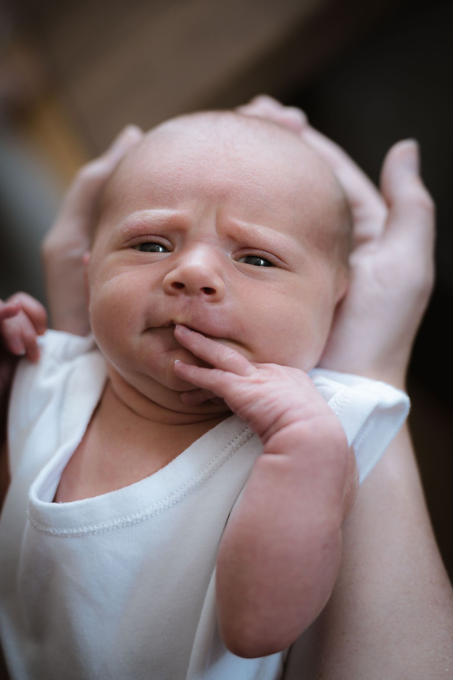 Close-up portrait of an infant in white clothing with hand near face.