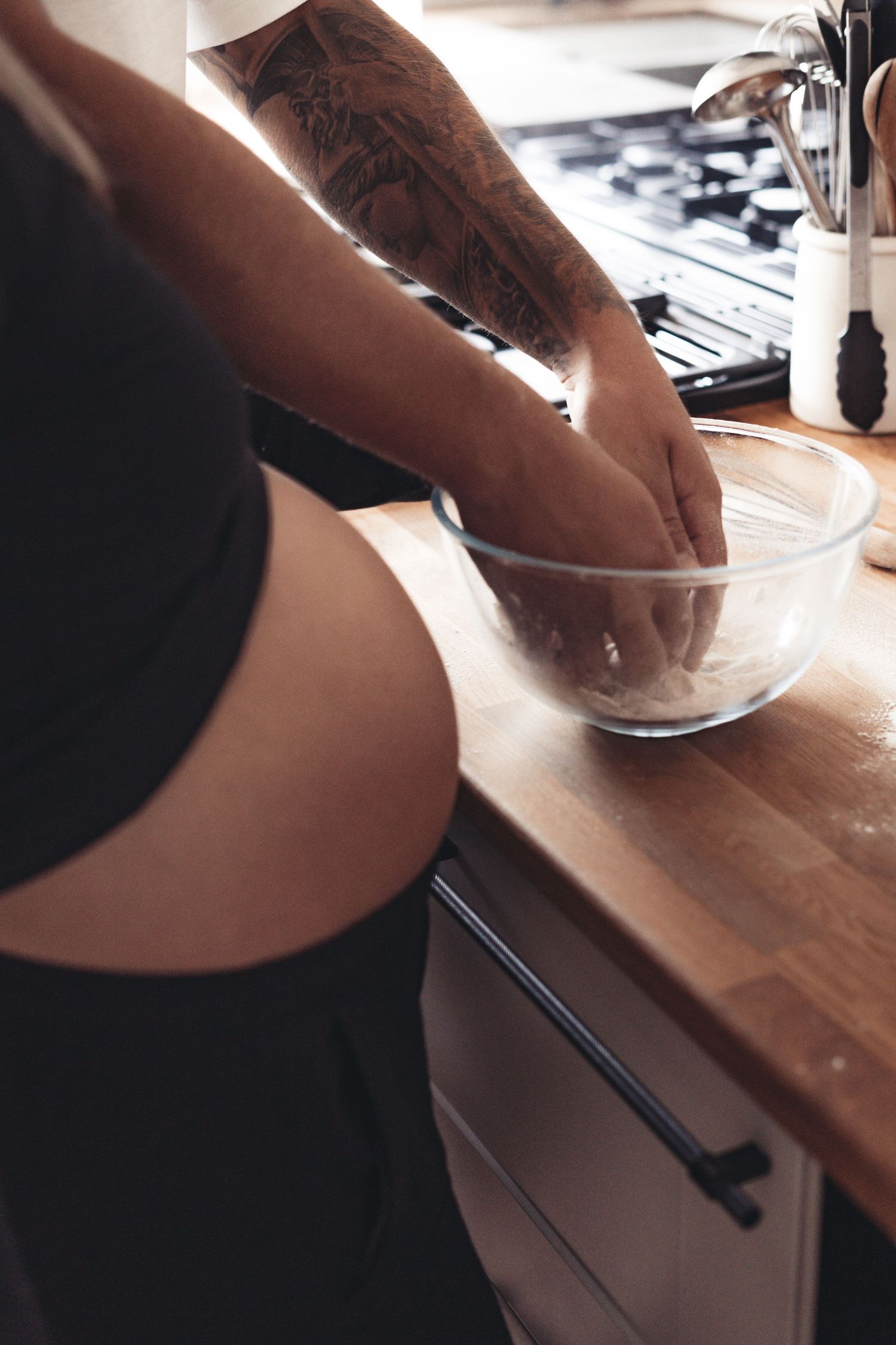 Tattooed arm mixing ingredients in glass bowl on wooden countertop.