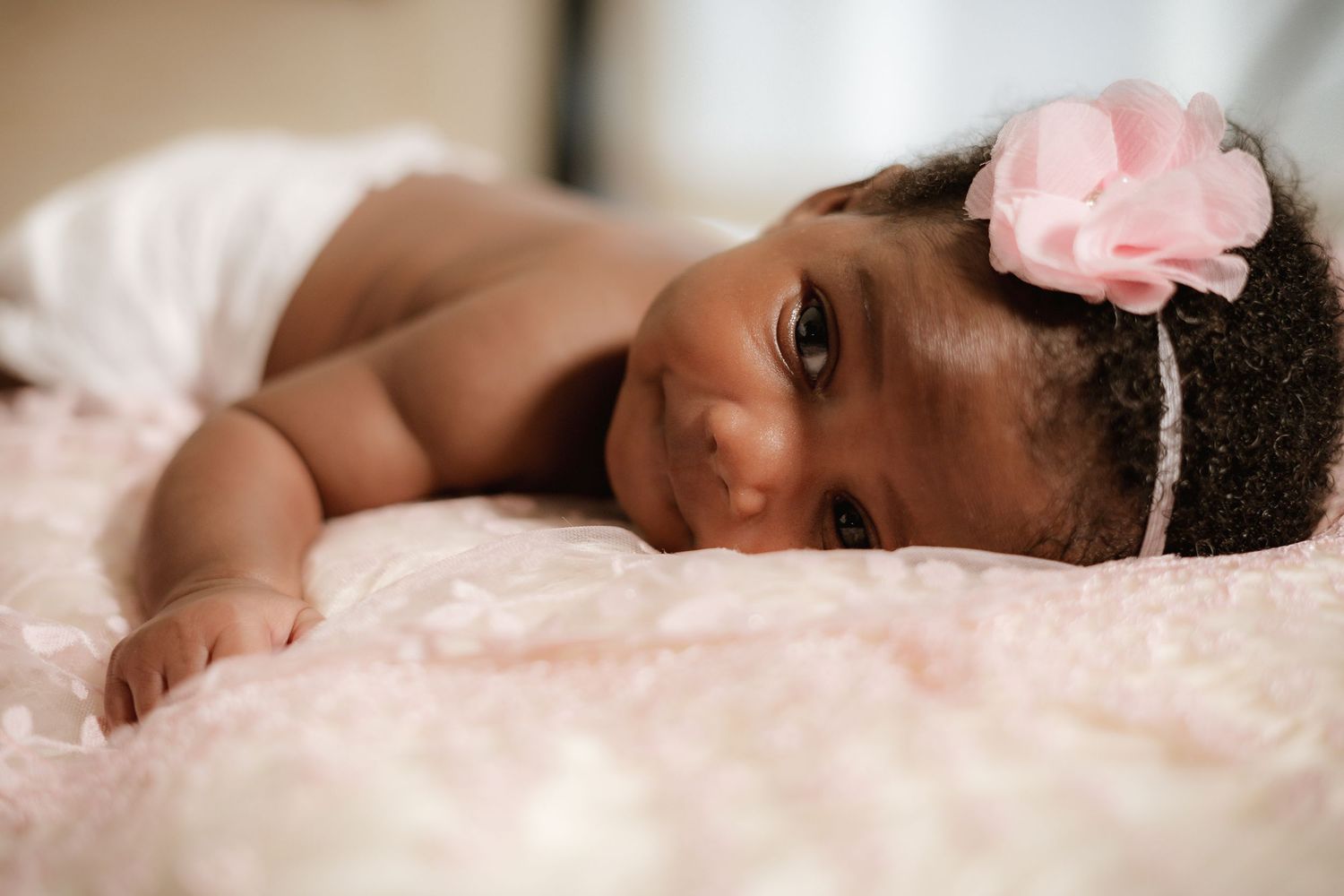 Baby wearing pink flower headband lying on blush pink bedding.