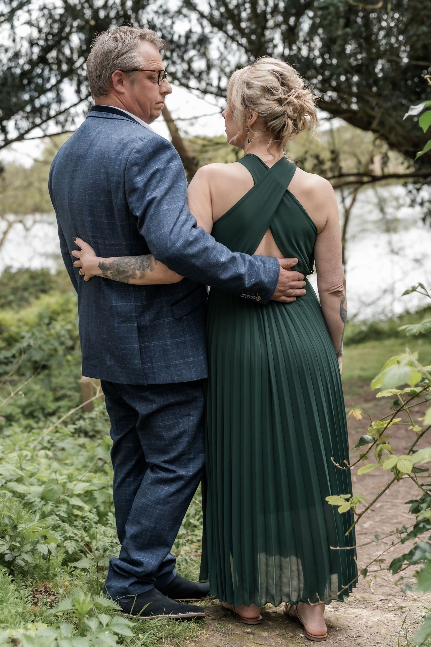 A couple embraces outdoors, with person wearing a blue suit and companion in an elegant green pleated dress.