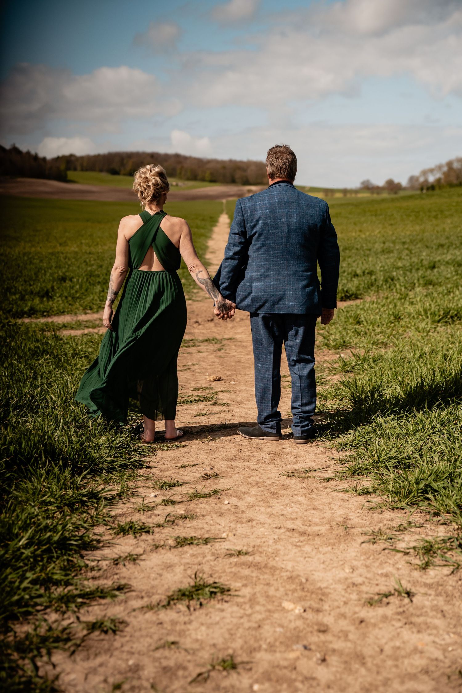 A couple holding hands while walking down a dirt path through green countryside on a cloudy day.