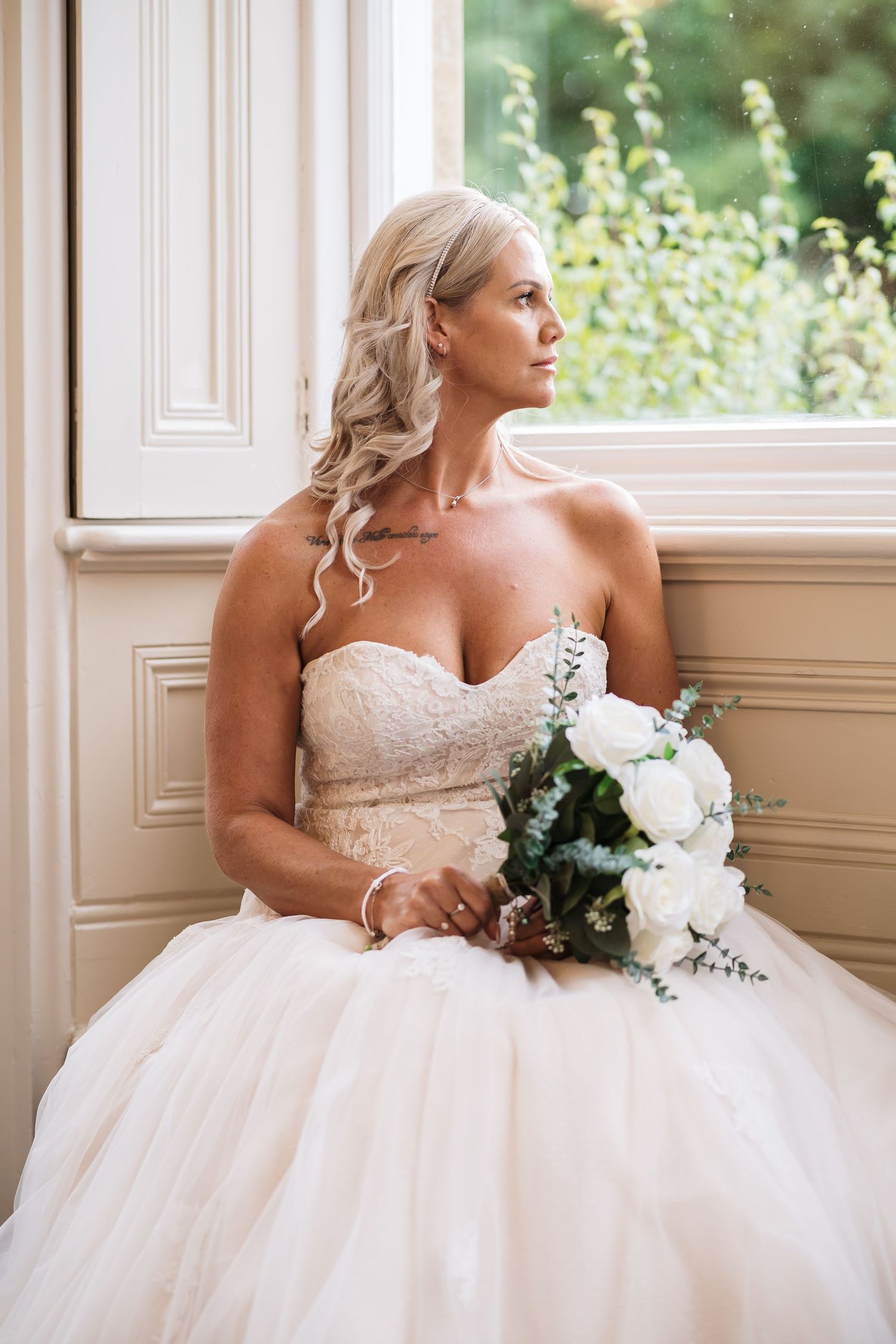 A bride in a white strapless ballgown wedding dress poses by a window holding a white floral bouquet.