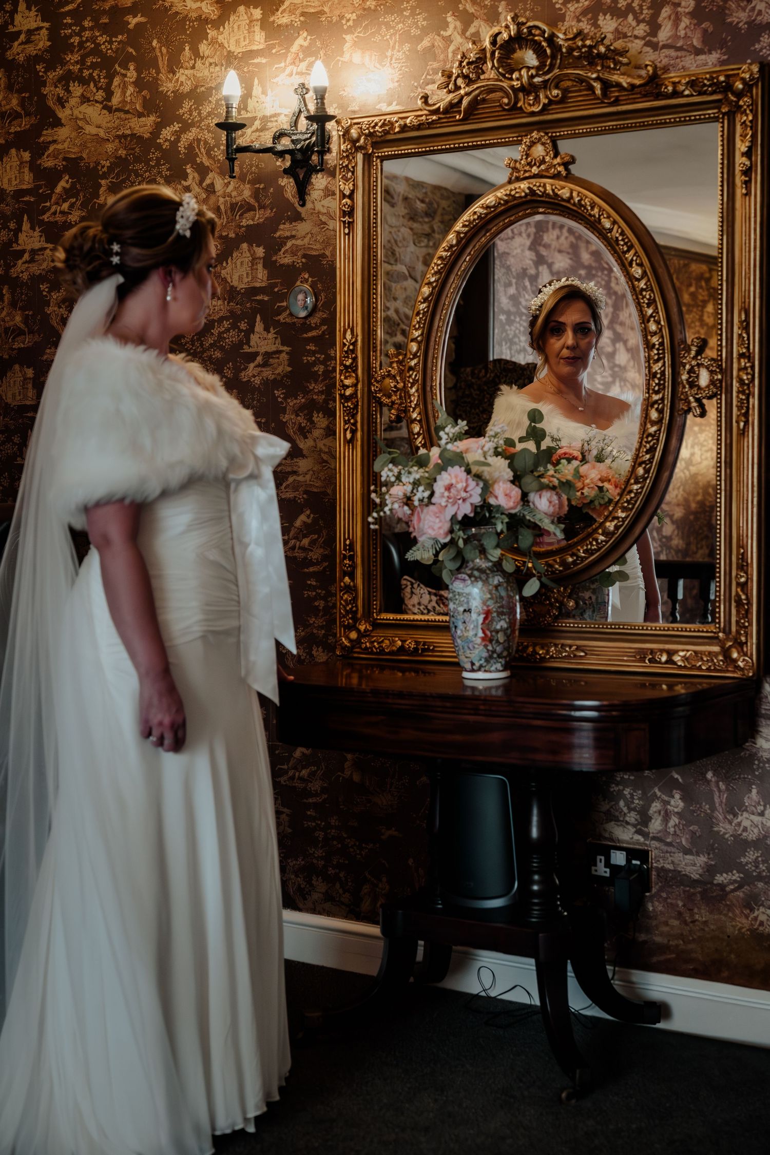 A bride in a winter wedding dress with fur stole gazes into an ornate vintage mirror.