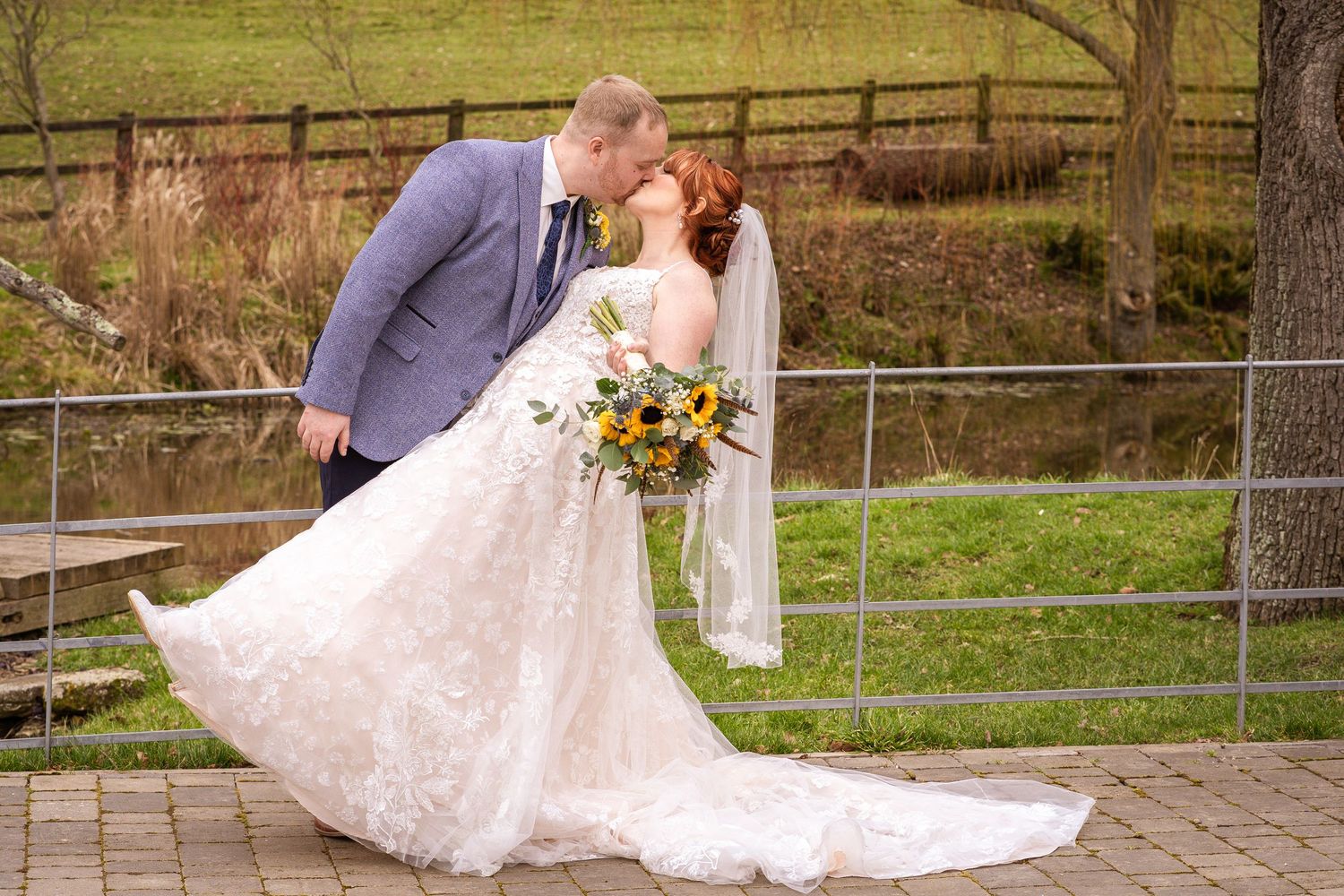 A romantic wedding couple sharing a kiss in front of a rustic wooden fence with a beautiful sunflower bouquet.