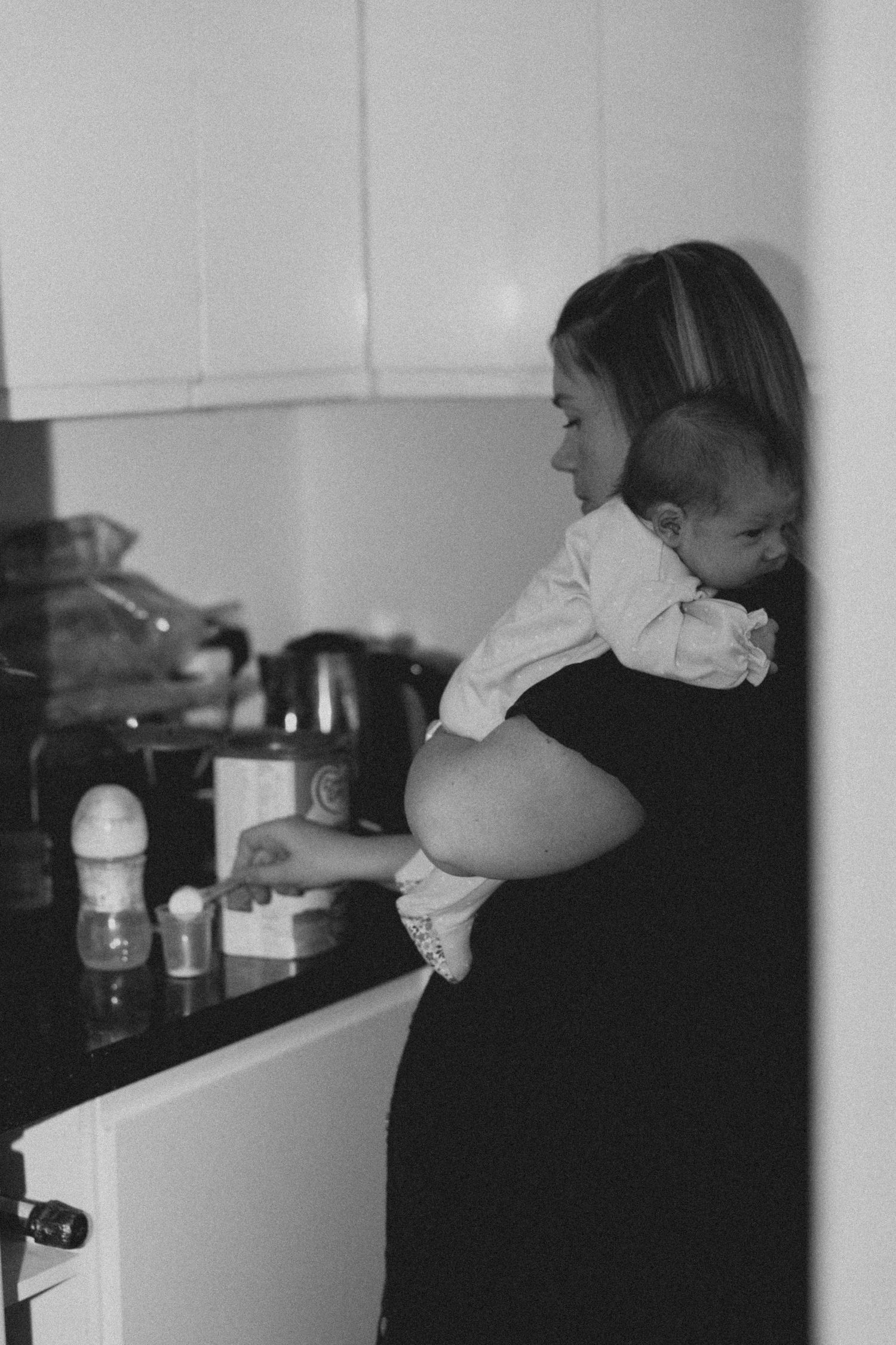 Side view of a pregnant woman in a kitchen holding a baby against her shoulder.