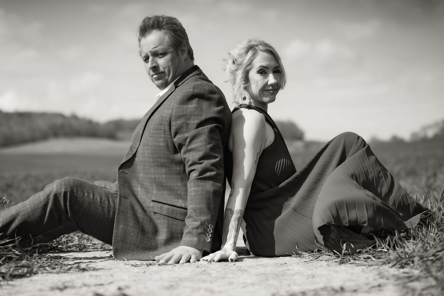 Stylish black and white portrait of a couple sitting back-to-back outdoors in a field with soft lighting and vintage mood.