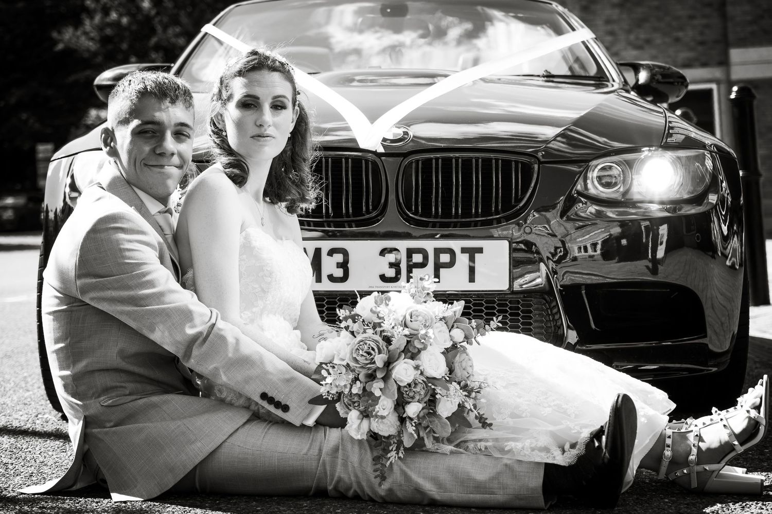 A black and white wedding photo with a couple sitting in front of a BMW luxury car.