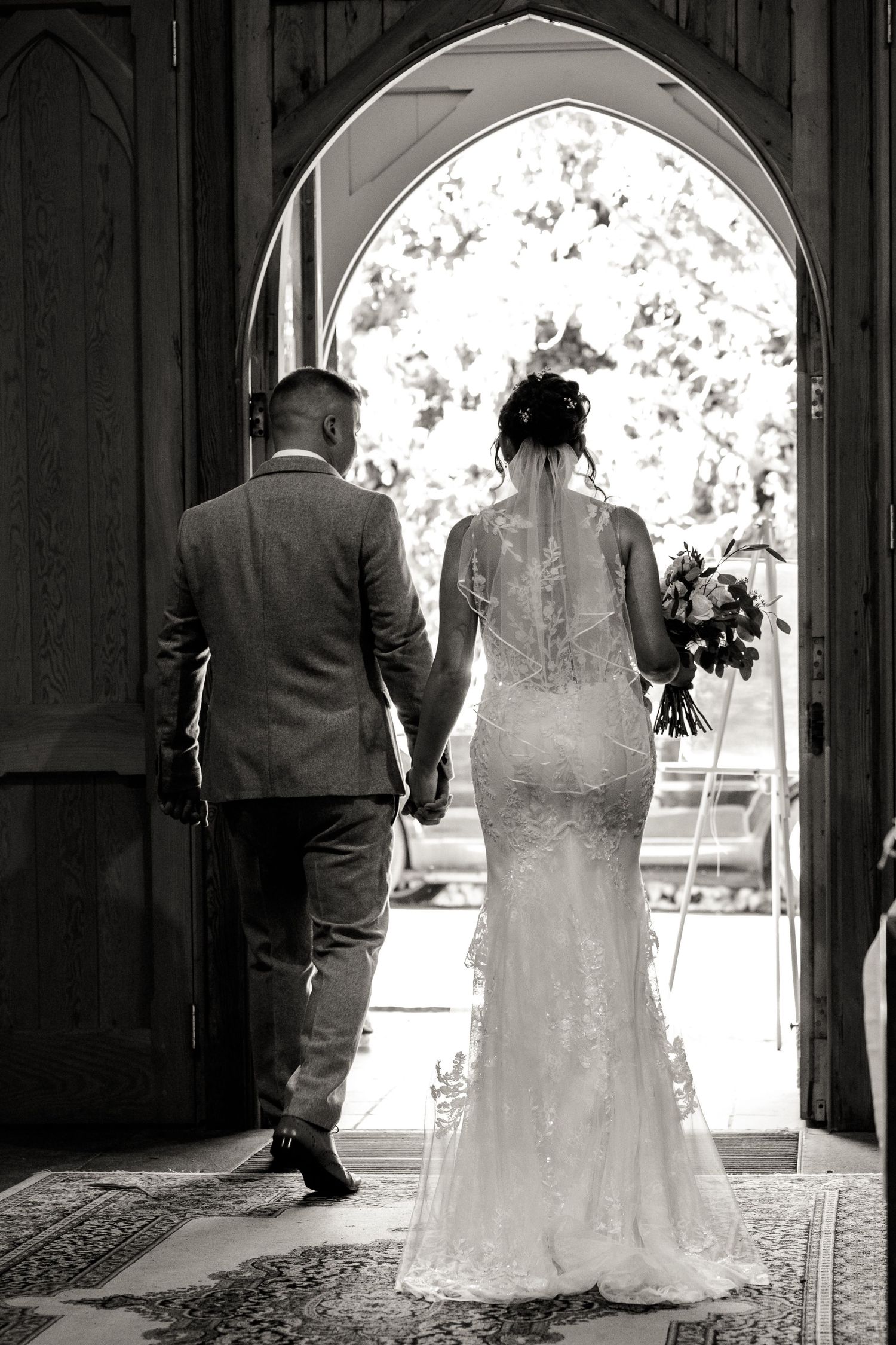 A black and white wedding photo showing a couple walking through an arched church doorway.