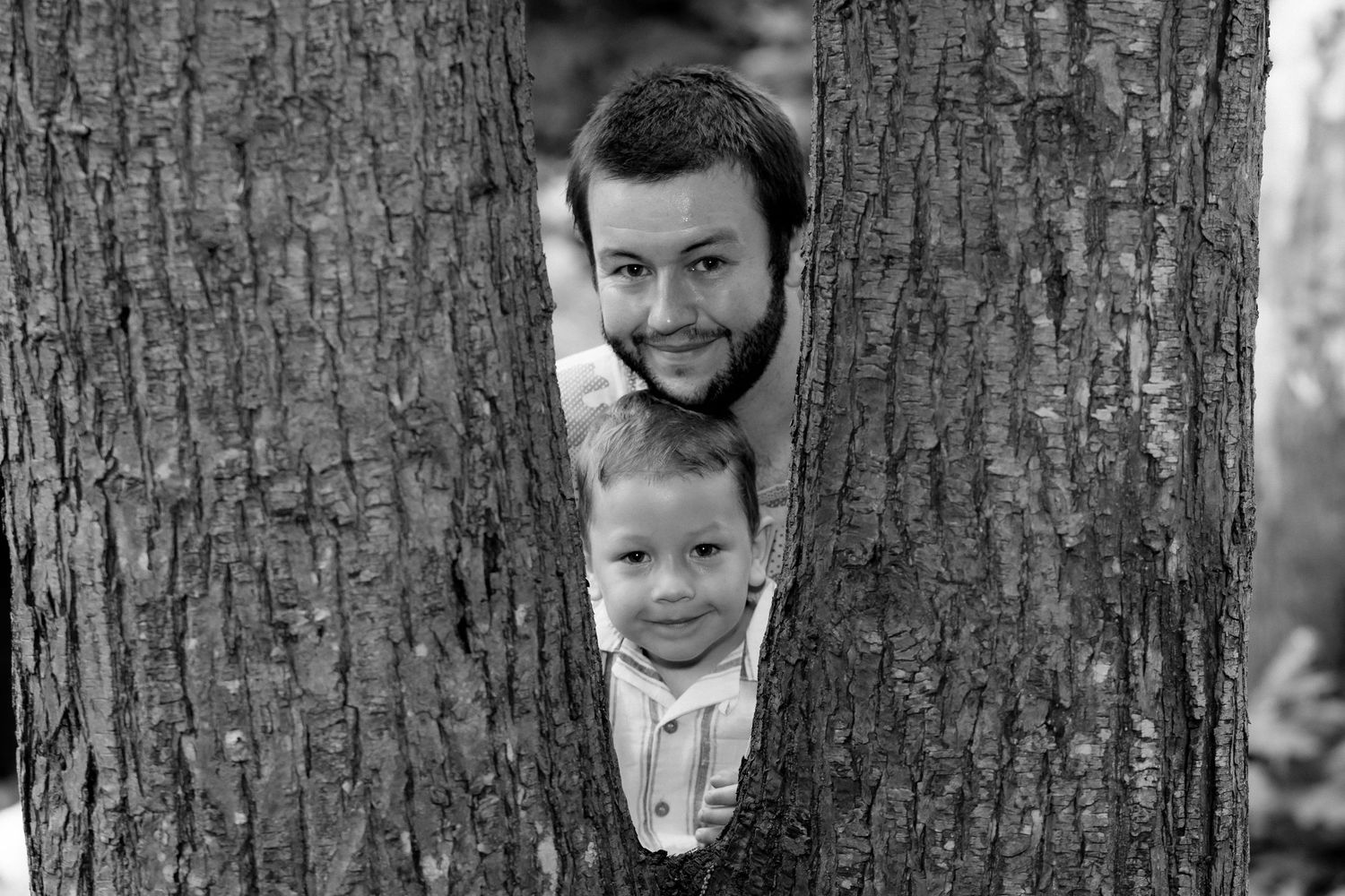 Black and white portrait of two people peeking between tree trunks in the woods.