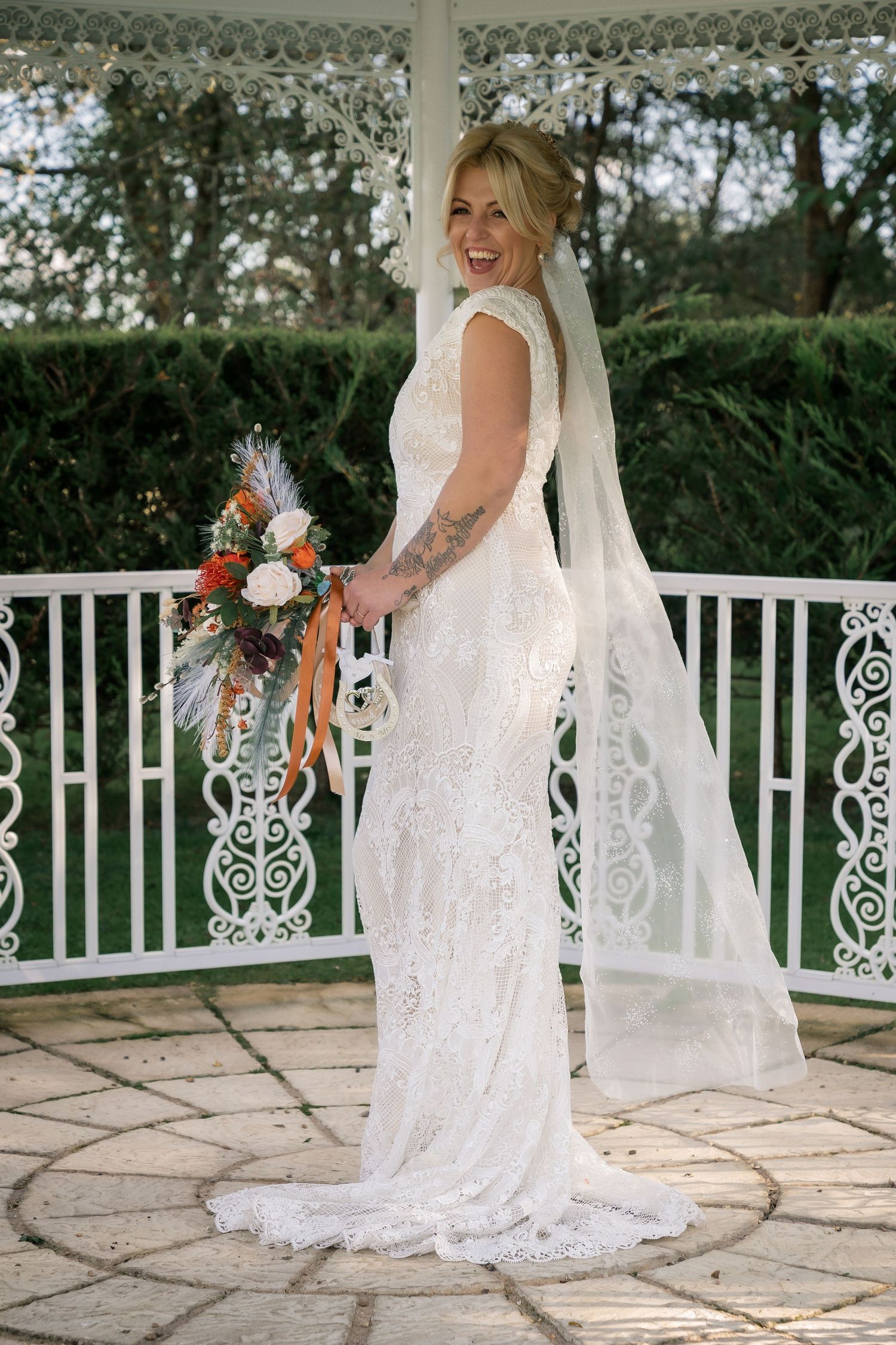 Joyful bride in lace dress with flowing veil poses on ornate white gazebo.