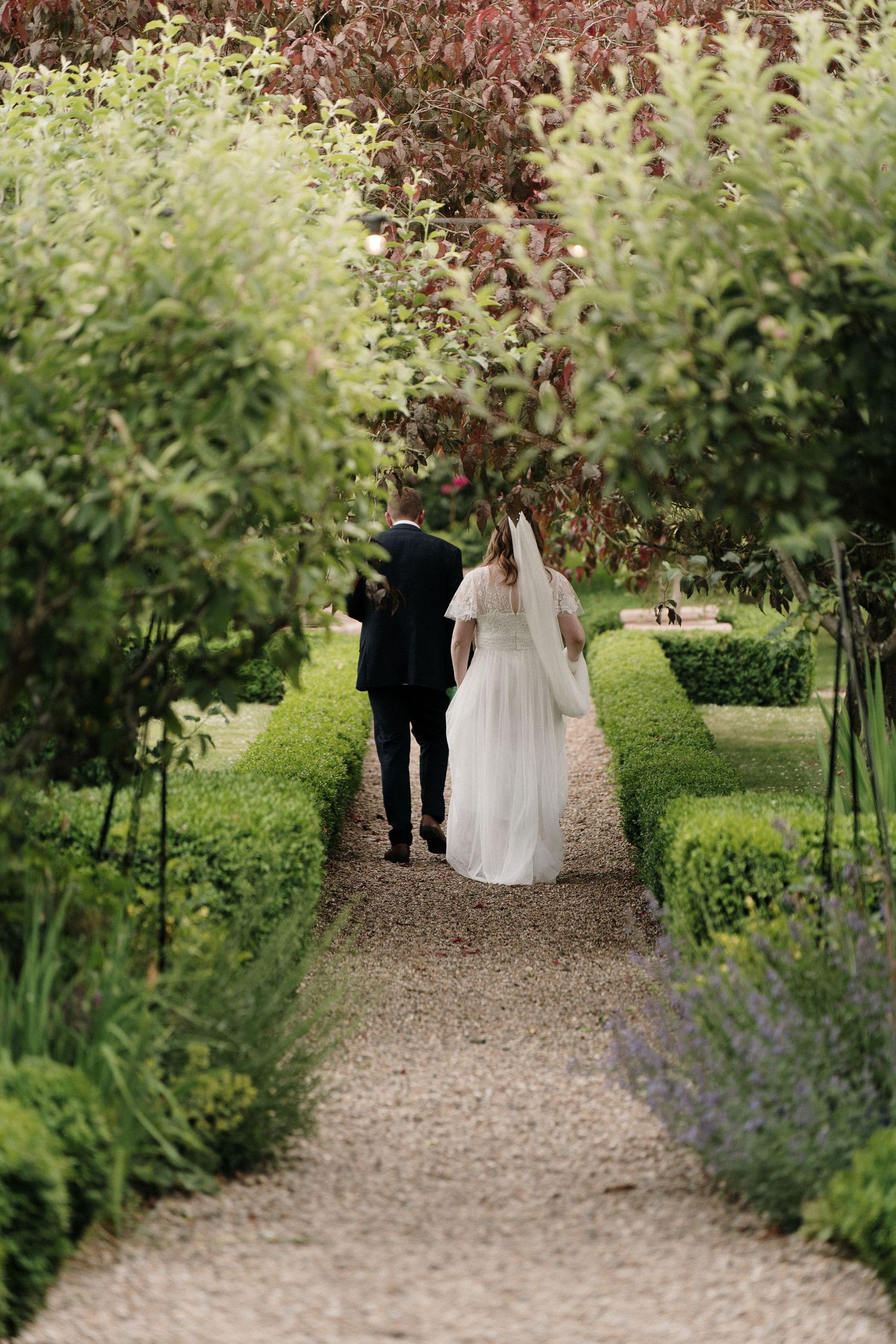 A couple walks down a garden path lined with lush greenery and flowers.