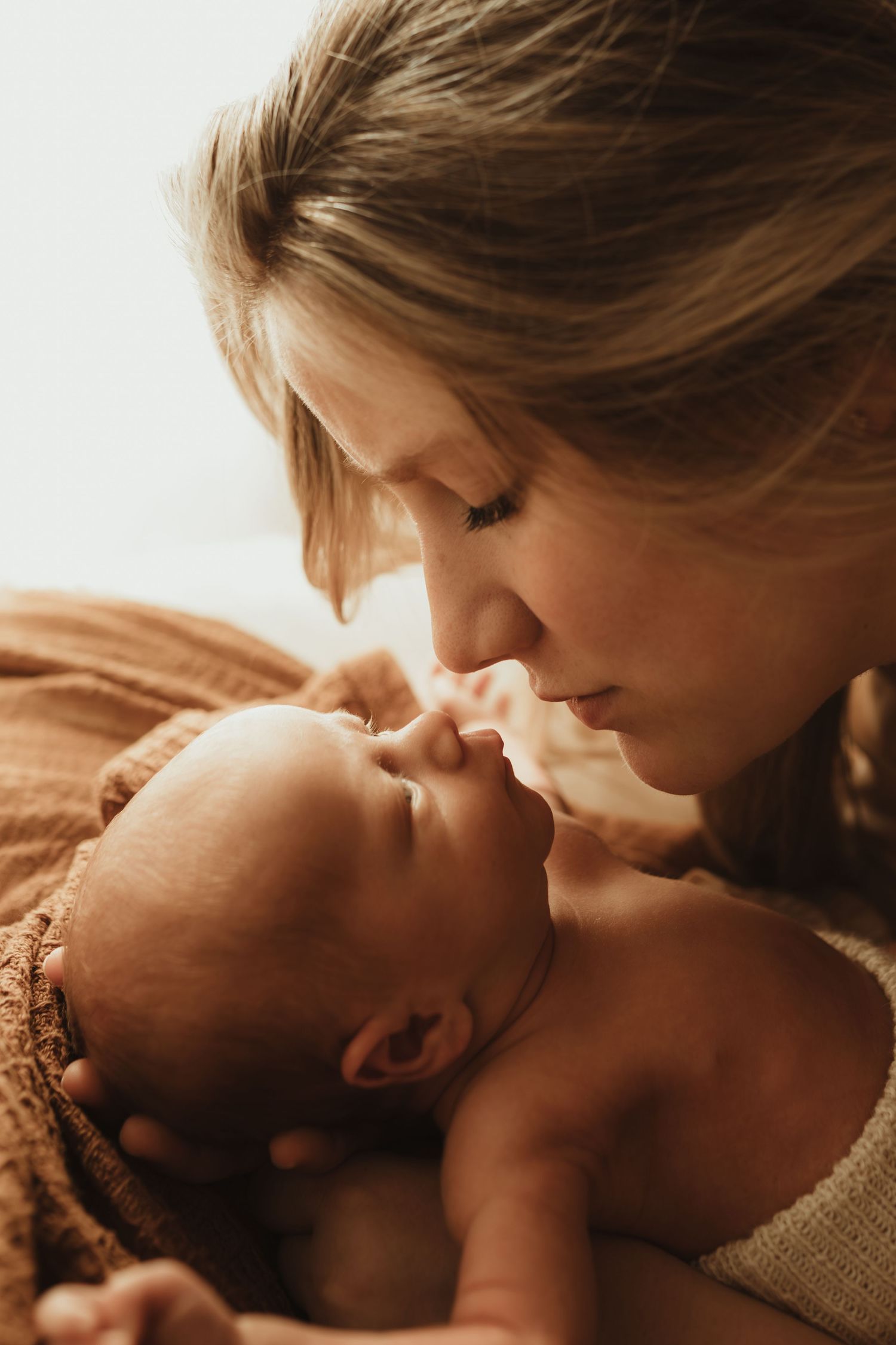 A tender moment as a young mother gently kisses her sleeping newborn baby wrapped in a warm brown blanket.