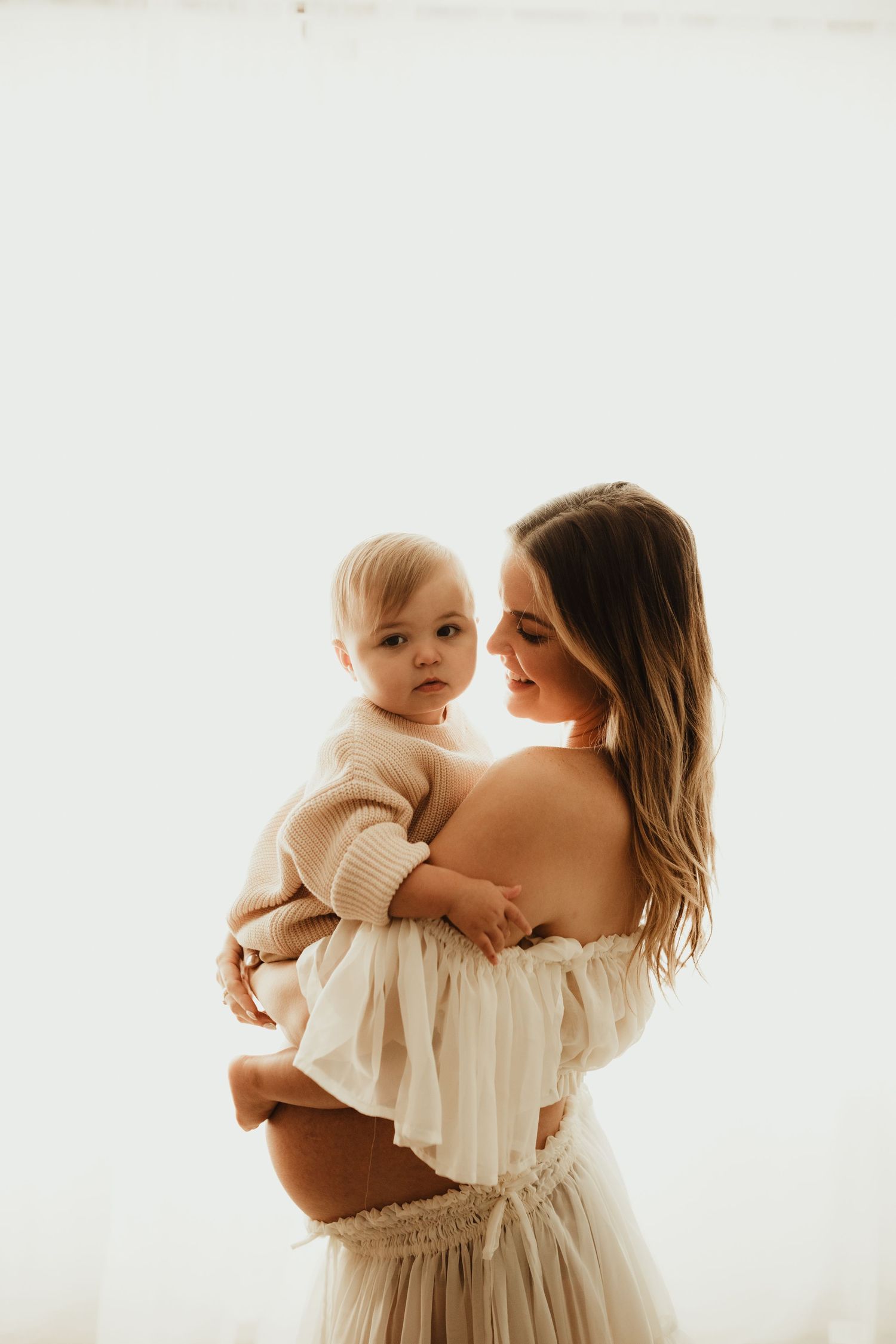 A family portrait series featuring tender moments between mother and baby in soft white clothing against a light backdrop.