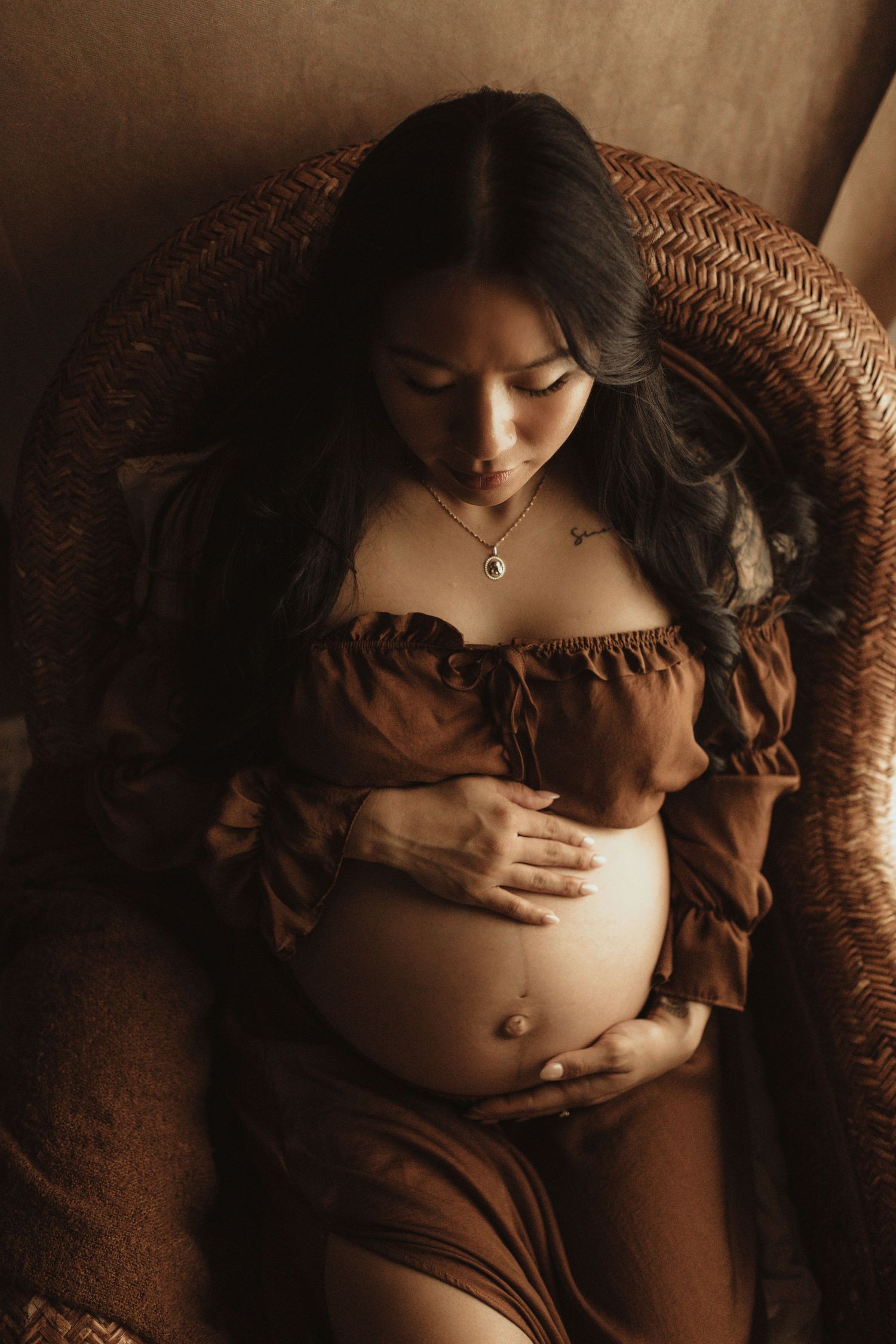 Pregnant woman in brown off-shoulder dress cradling her baby bump while seated in a wicker chair.