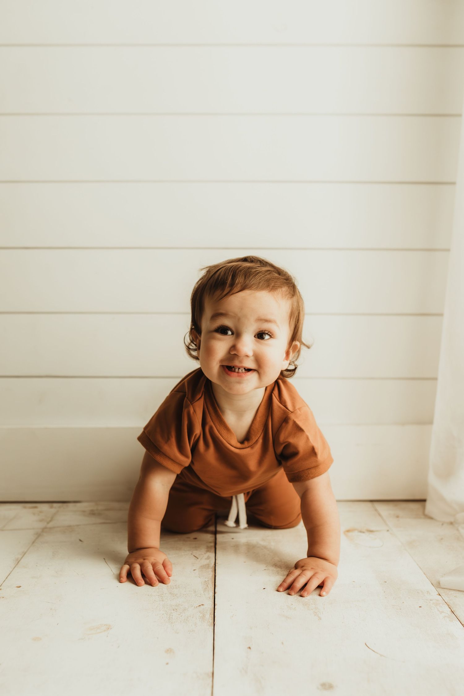 Baby in brown outfit crawls on white wooden floor against shiplap wall background.