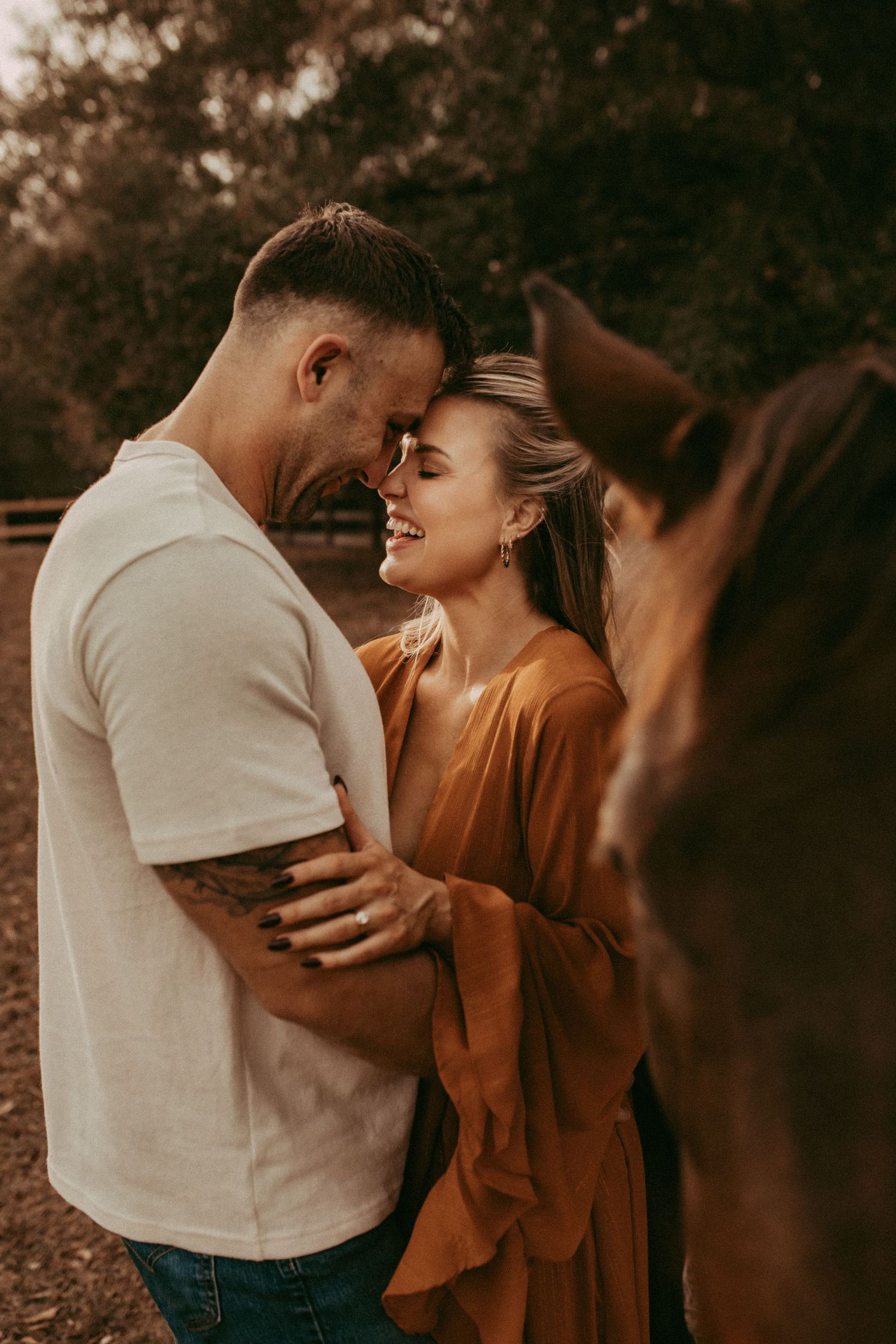 A couple shares a romantic moment outdoors near a horse, the woman smiling in an amber dress while the man wears a white tee.
