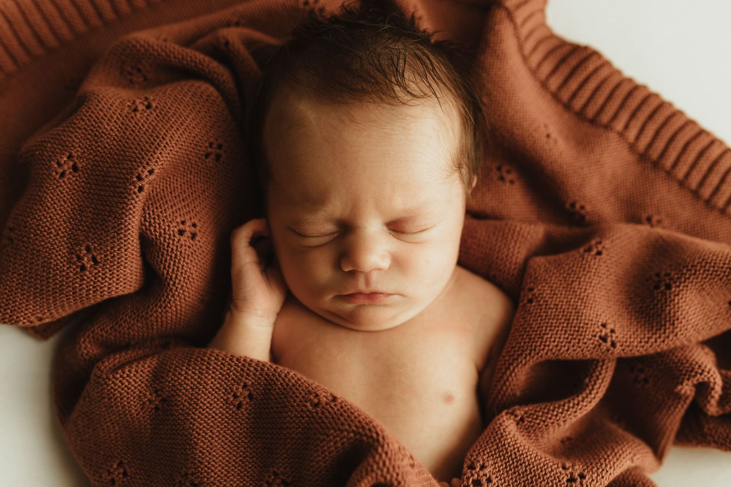 Peaceful newborn baby sleeping wrapped in a rustic terracotta knit blanket in a cozy posed newborn photography setup.