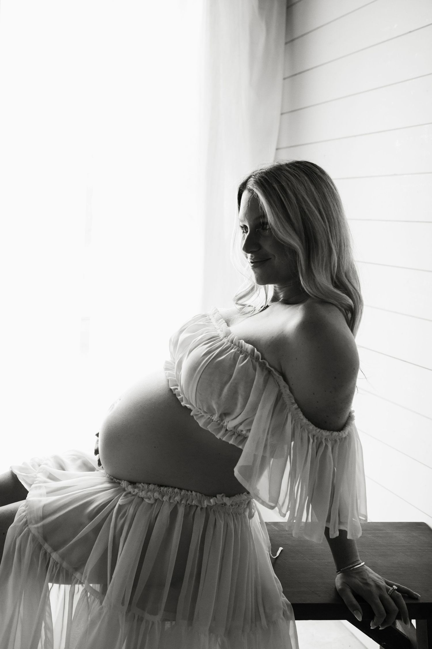 Black and white maternity photo of pregnant woman in sheer dress posing by bright window, cradling baby bump.