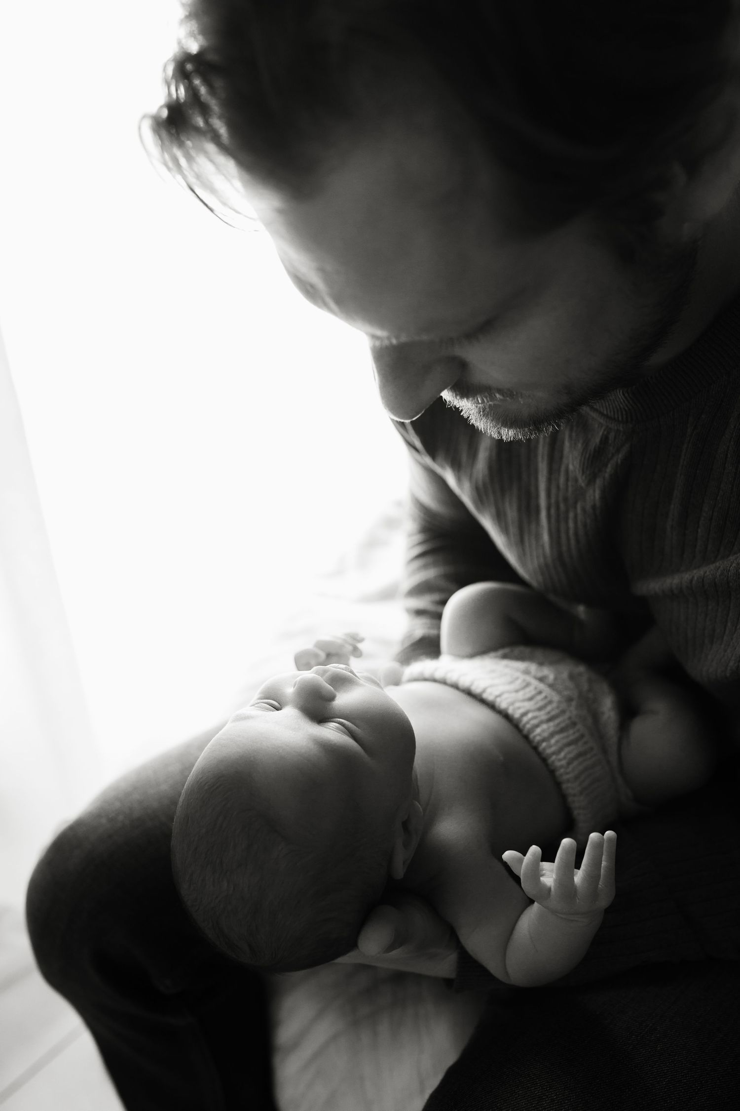 Black and white photo of a father tenderly cradling his newborn baby near a bright window.
