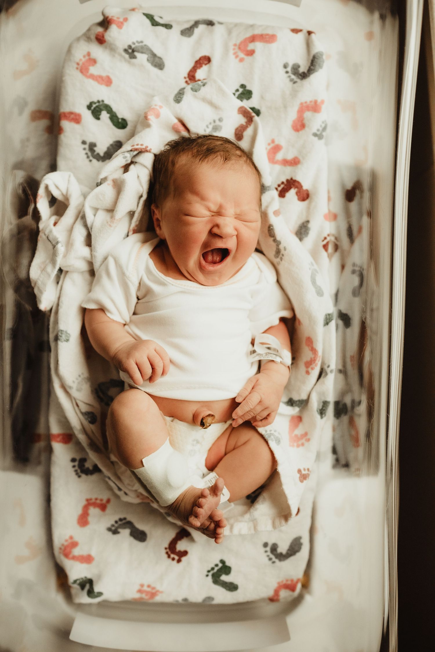 Newborn baby yawning in hospital bassinet with colorful footprint blanket, wearing white onesie.